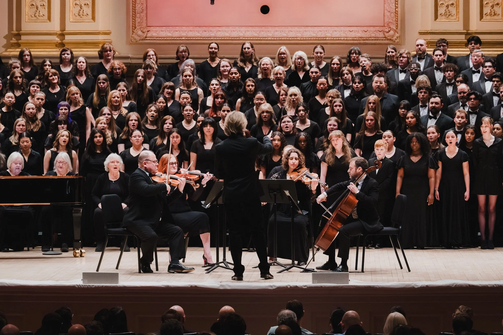 Conductor with back to camera leads a string quartet in front of a massive combined choir on risers at Carnegie Hall Stern Auditorium, salmon-toned panels visible behind the ensemble