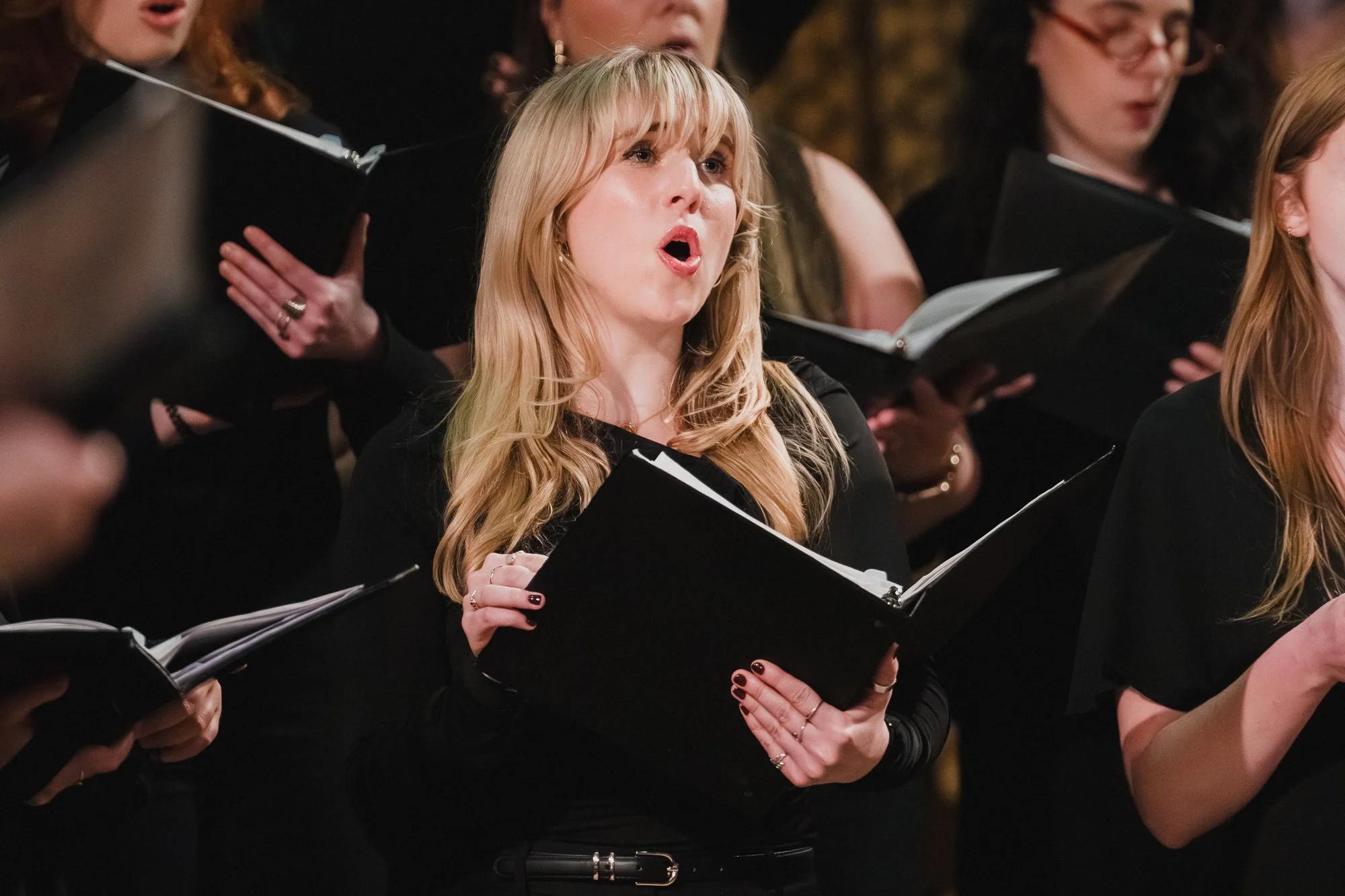 Blonde soprano in black turtleneck singing with folder open, surrounded by other singers in soft focus.