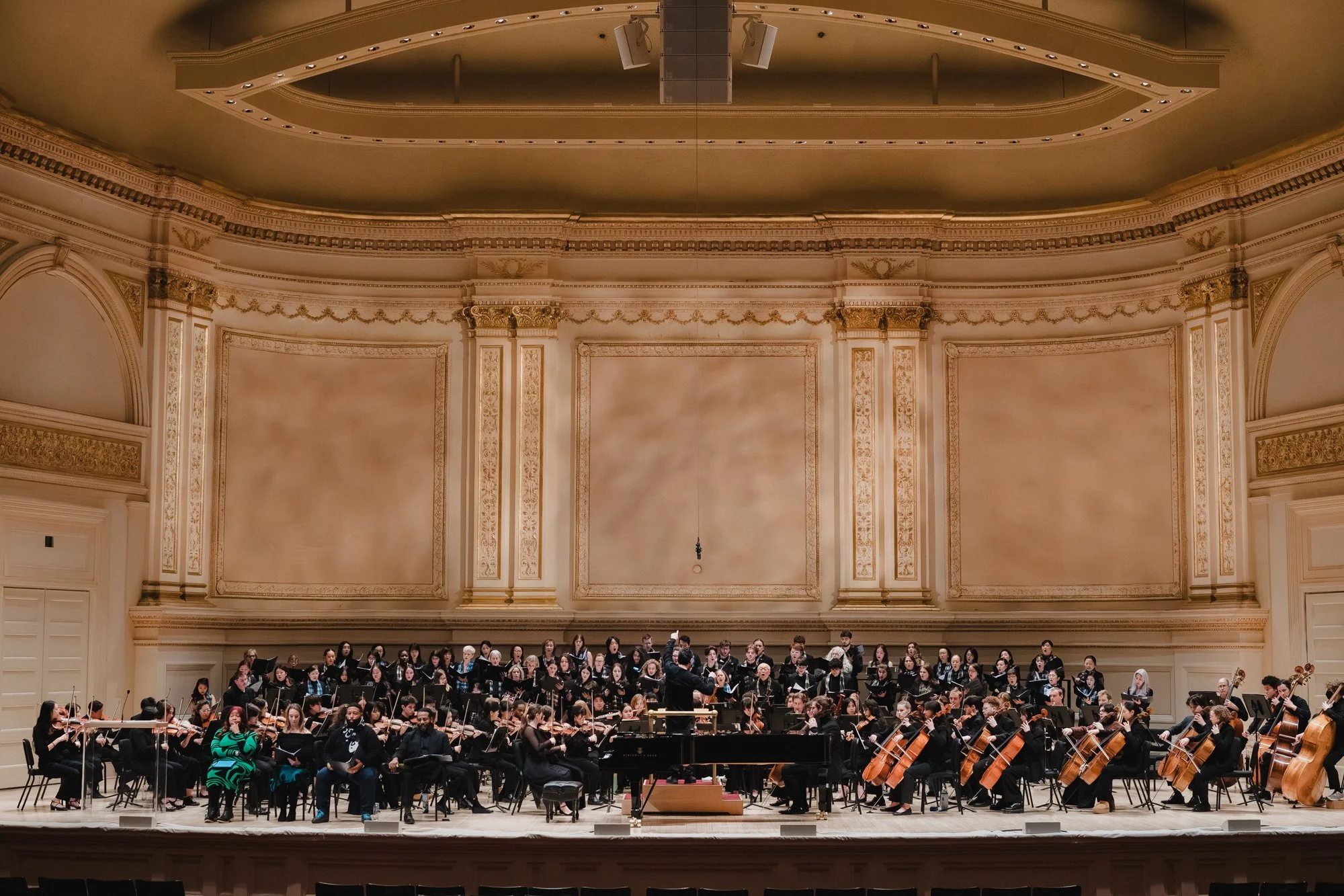 Full orchestra and chorus fill the stage at Carnegie Hall's Stern Auditorium, ornate cream and gold walls and ceiling visible above.