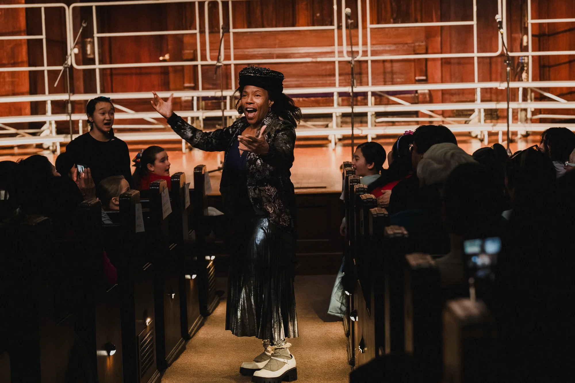 Song leader in metallic skirt and patterned jacket leading singalong from the aisle with arms raised as audience members follow along at the New York Society for Ethical Culture