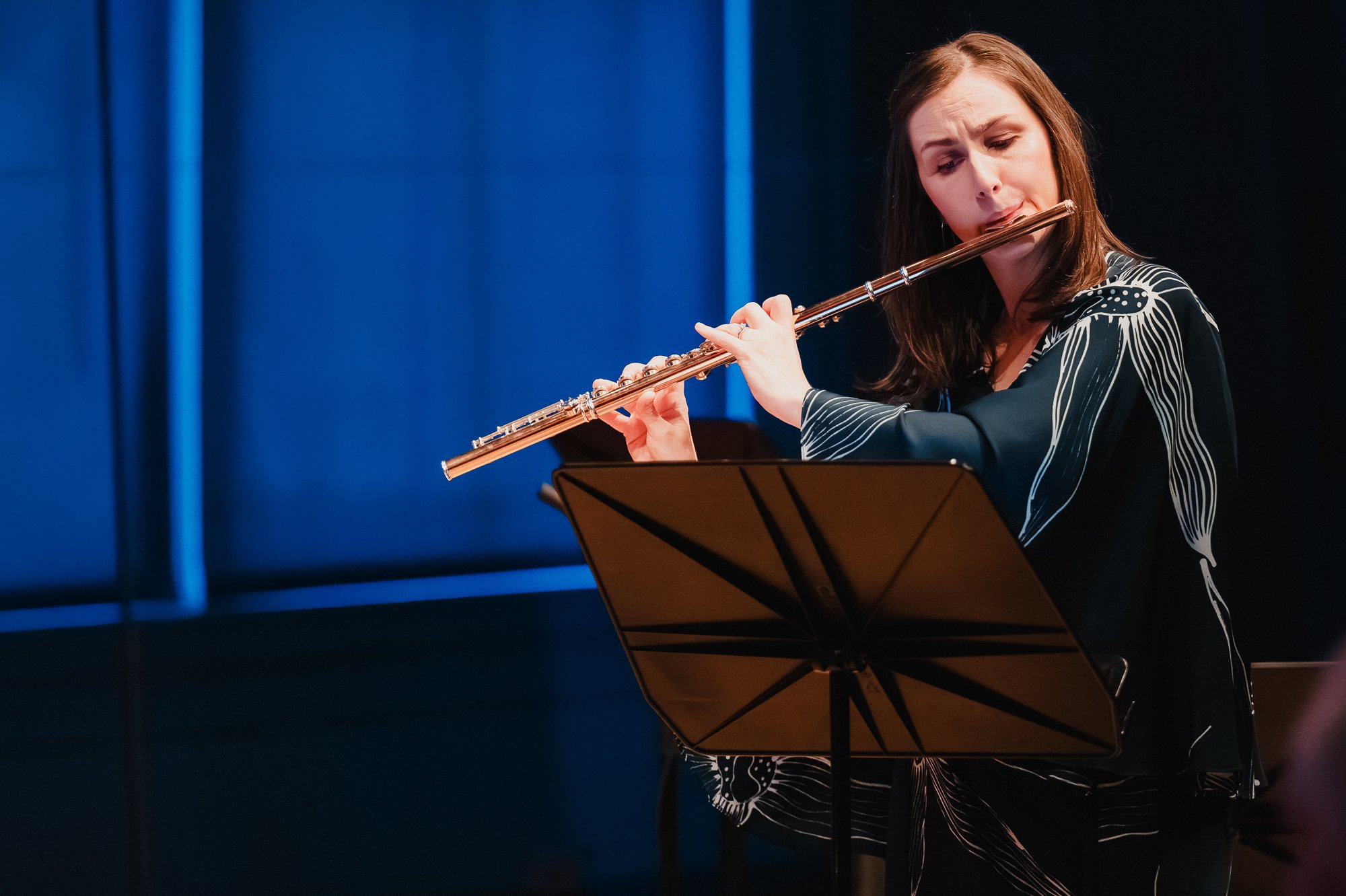 A flutist with dark hair plays against a deep blue background, eyes focused downward on a music stand, wearing a dark patterned blouse.