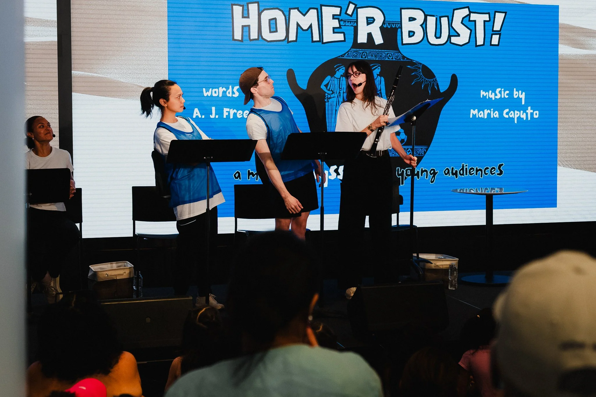 Three performers on a low stage, one standing and holding a clarinet while speaking, two seated behind her at music stands, a 'Home'r Bust!' title card on a screen behind them, audience in the foreground