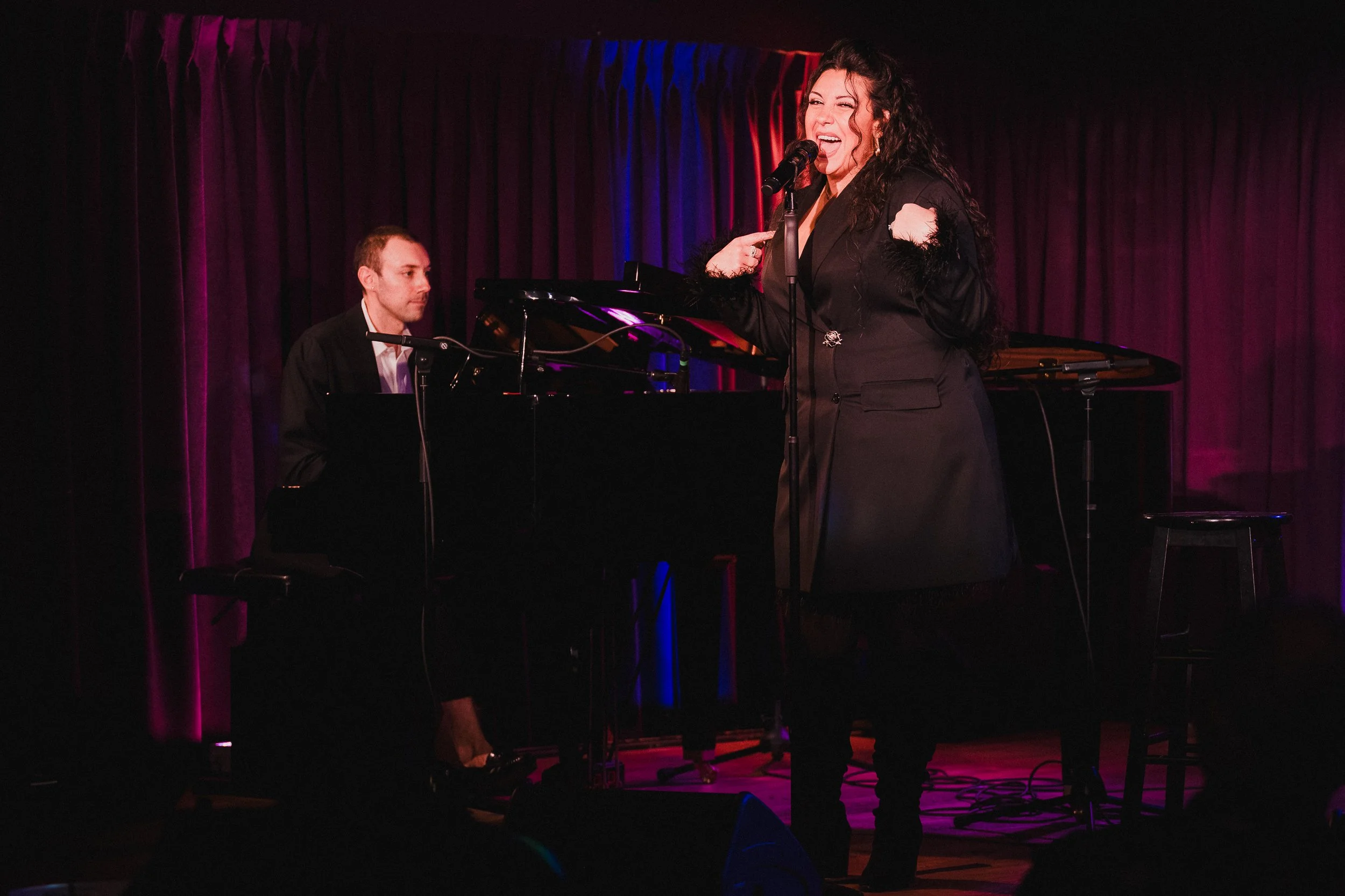Mary DiGangi singing at a standing microphone at The Green Room 42, Mike Pacifico at the piano to her left, red and blue curtain behind them