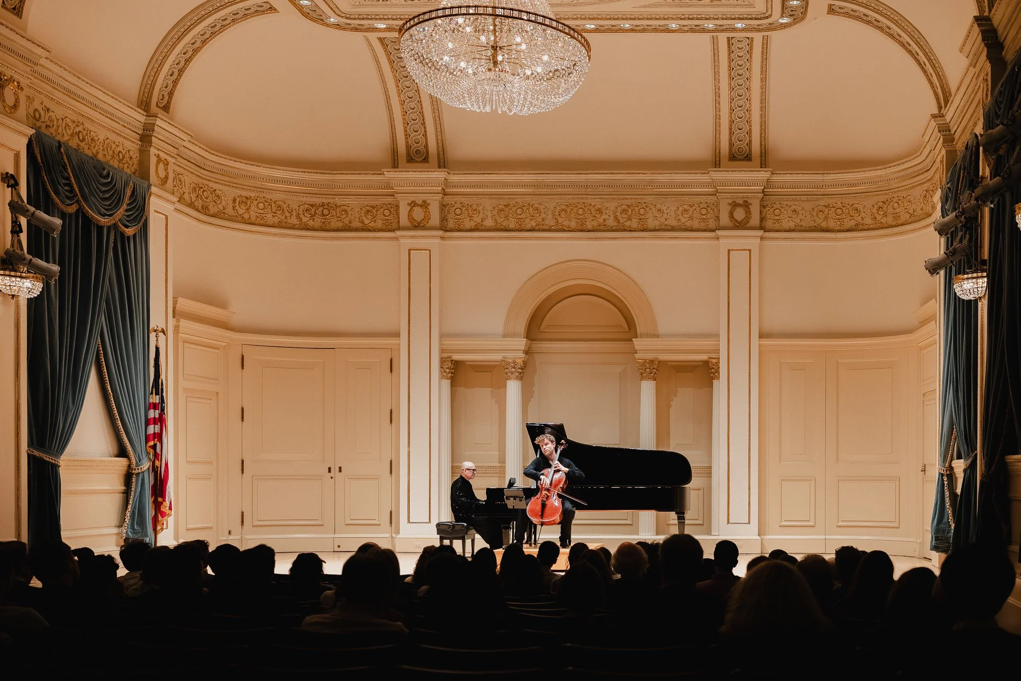 Wide view of Weill Recital Hall from the back of the house, silhouetted          audience watching Rainer Crosett on cello and Victor Santiago Asunción at the    Steinway under the crystal chandelier.