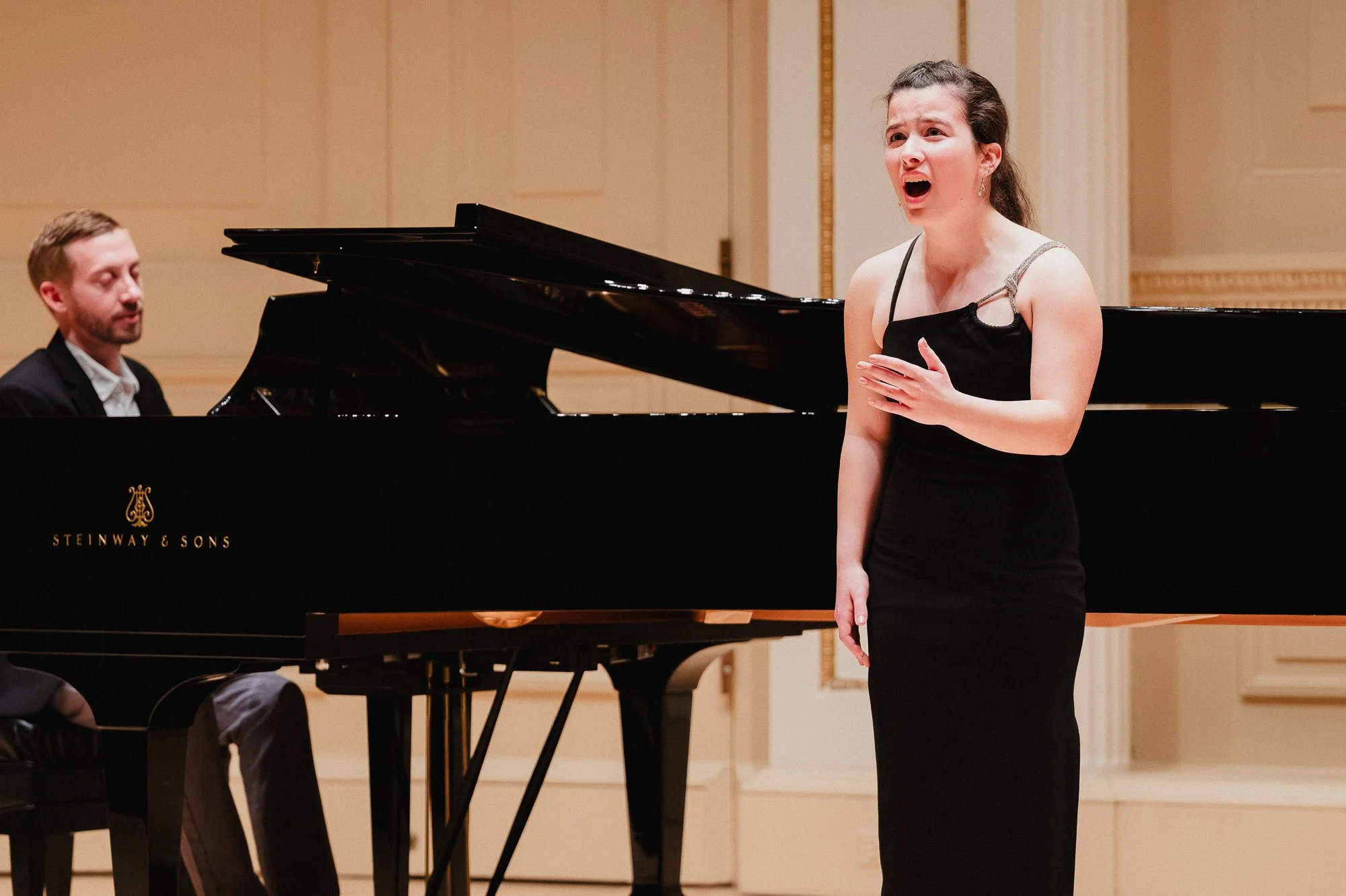 A young woman in a black gown sings beside a Steinway grand piano, one hand raised toward her chest, mouth open mid-phrase, man at piano visible in soft focus behind