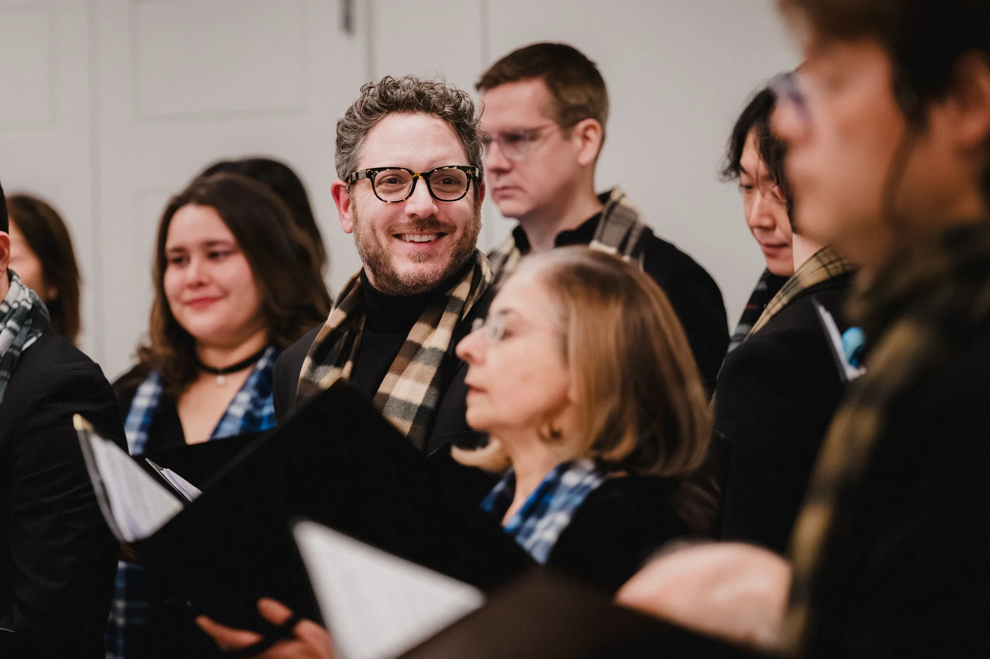 Male singer in glasses and tan plaid scarf smiles during Carnegie Hall rehearsal, other choir members in soft focus around him.