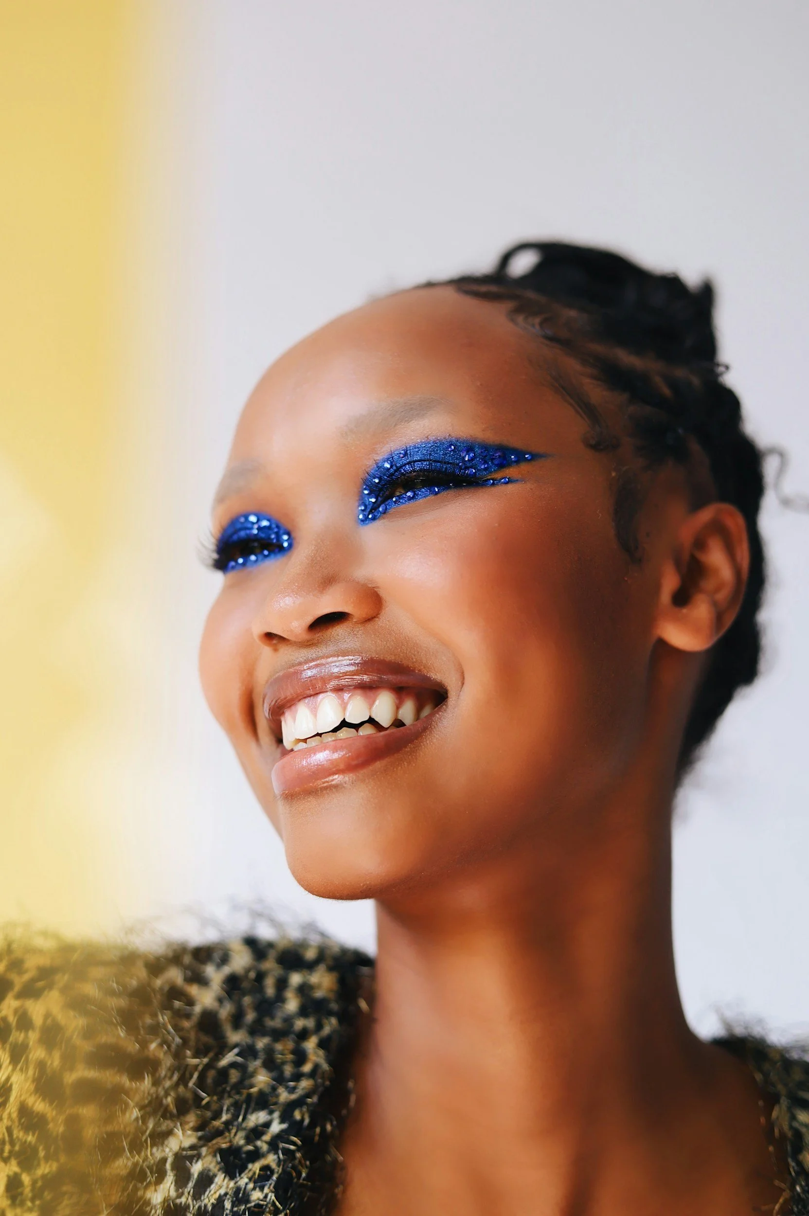 A woman with short curly hair and dark skin is smiling, with blue glittery eye makeup and a patterned dress.