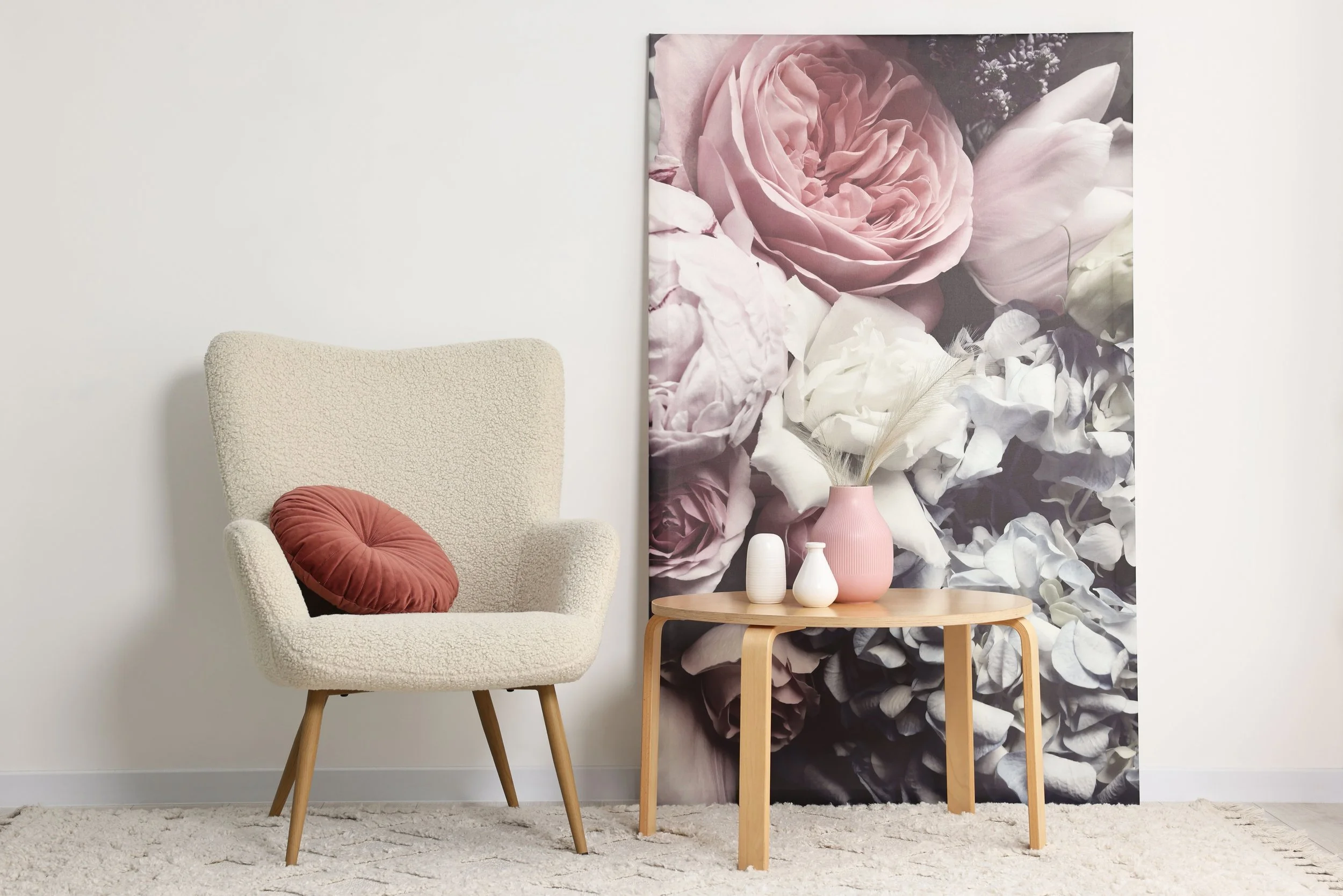 A cozy living room corner with a beige armchair, a small wooden side table holding pink and white vases with dried pampas grass, and a large floral artwork featuring pink, white, and lavender roses on the wall behind.