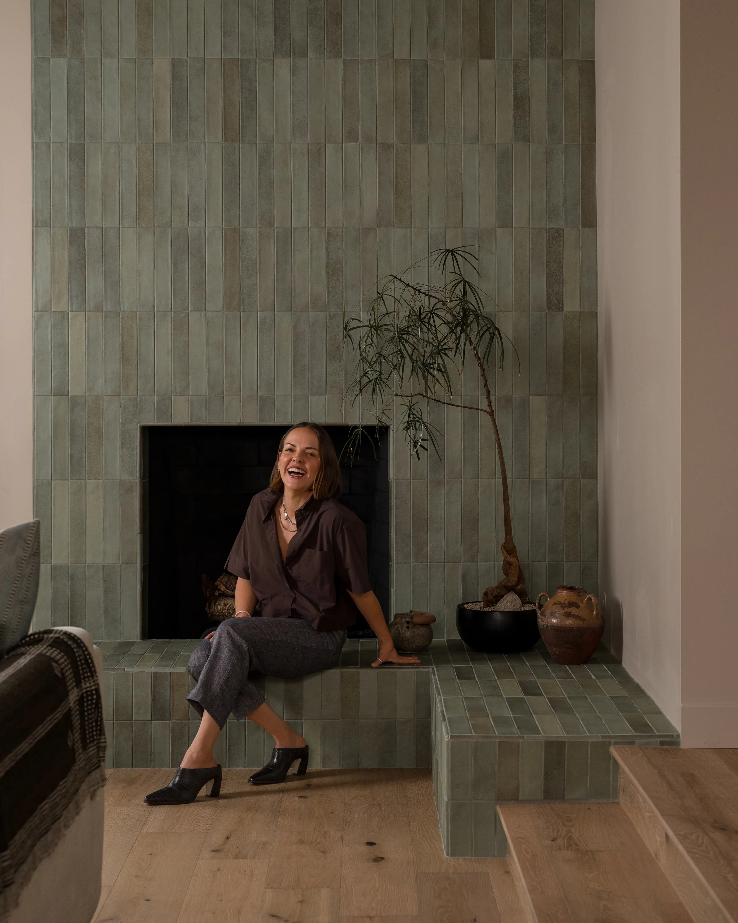 Heather KW Styles, Redondo Beach interior designer, is smiling and laughing while sitting on a green tiled fireplace hearth in a space she designed.
