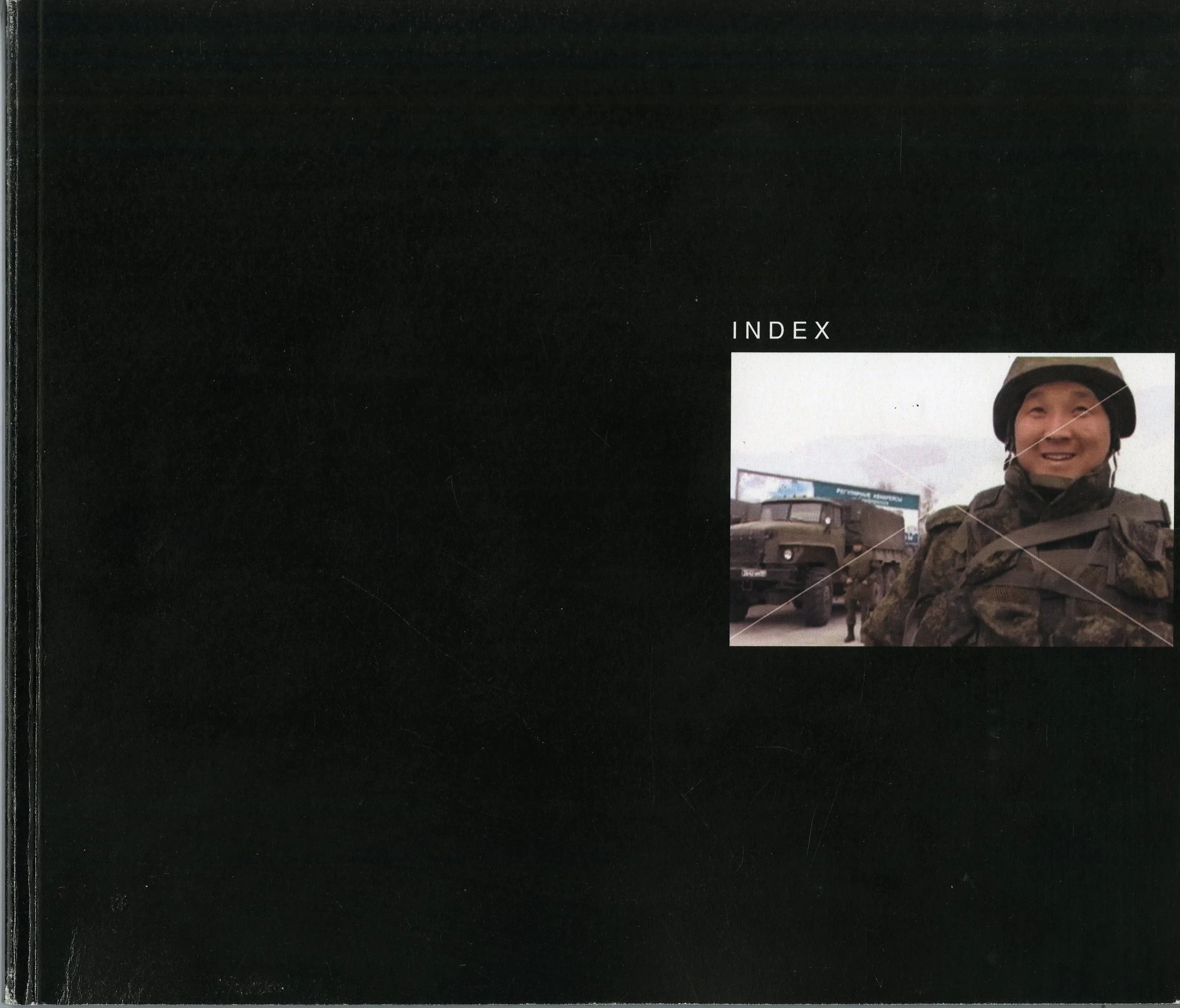 Alt Text: All-black book cover, interrupted only by small, white, all caps title reading "Index" just above small, color photograph of soldier smiling in foreground, standing in front of armored truck. A thin white "X" crosses through the image, remi