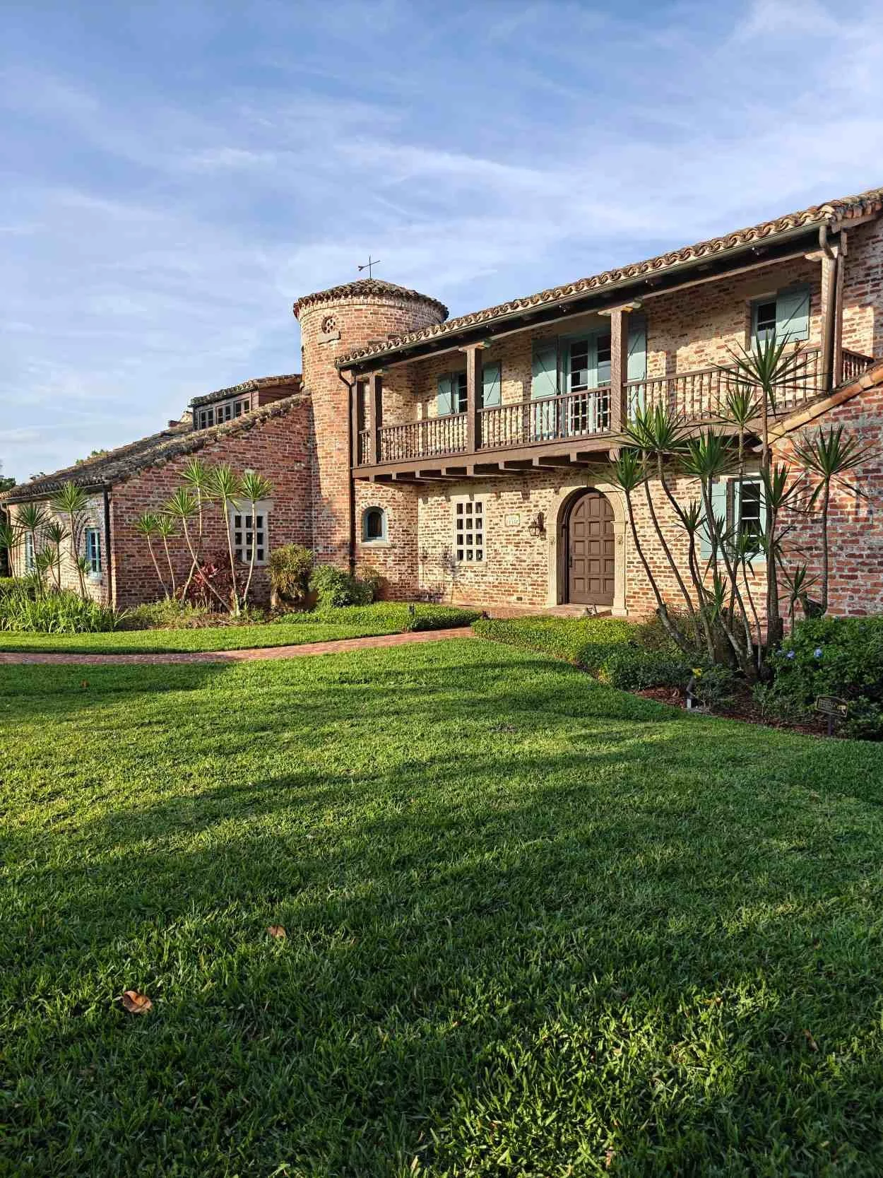 A brick house with a tower, balcony, and arched front door, surrounded by a well-maintained lawn and landscaping under a blue sky.