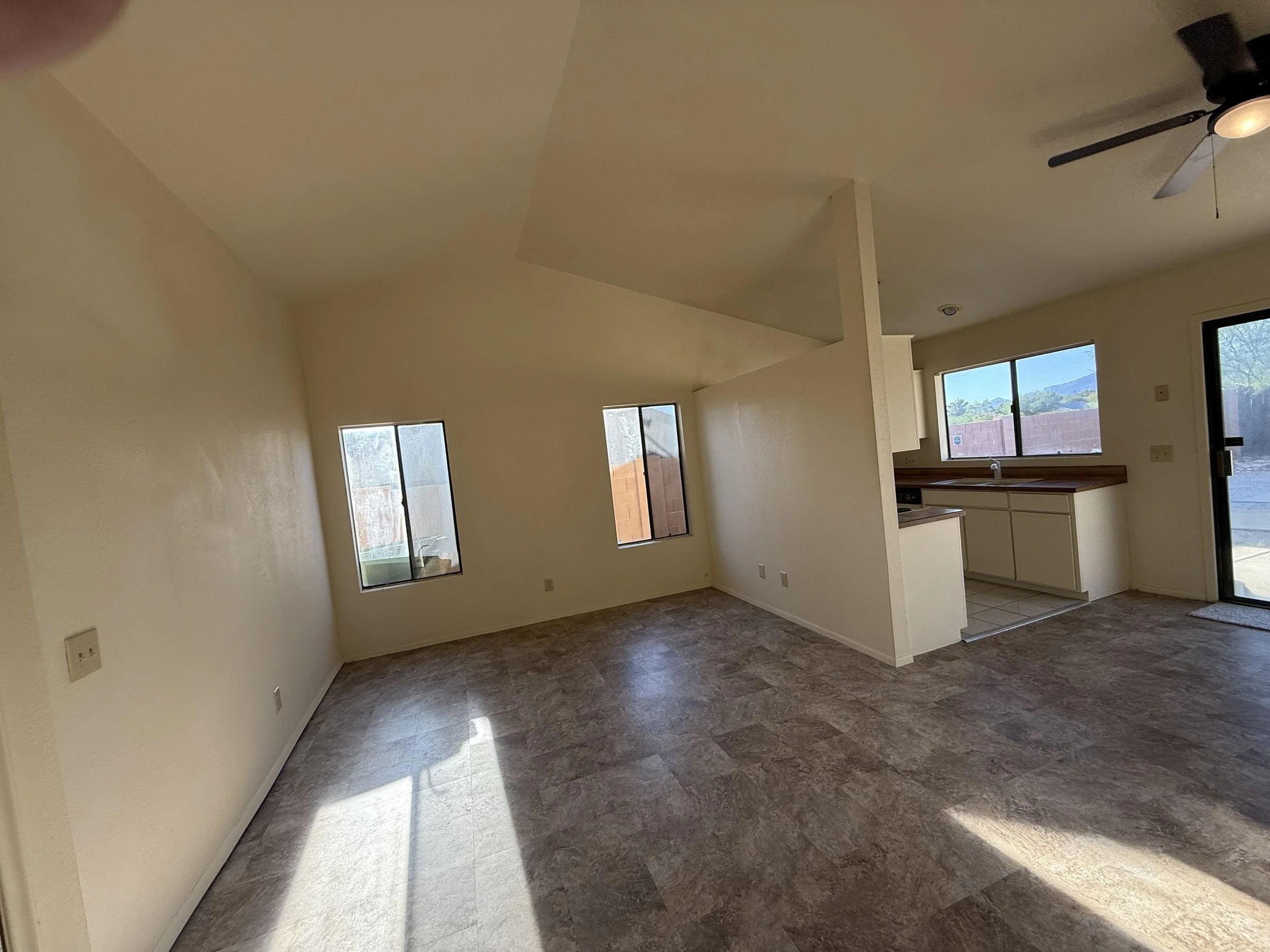 Empty living room with beige walls, tiled floor, and open kitchen with white cabinets, wooden countertop, and large windows, in a sunny house.