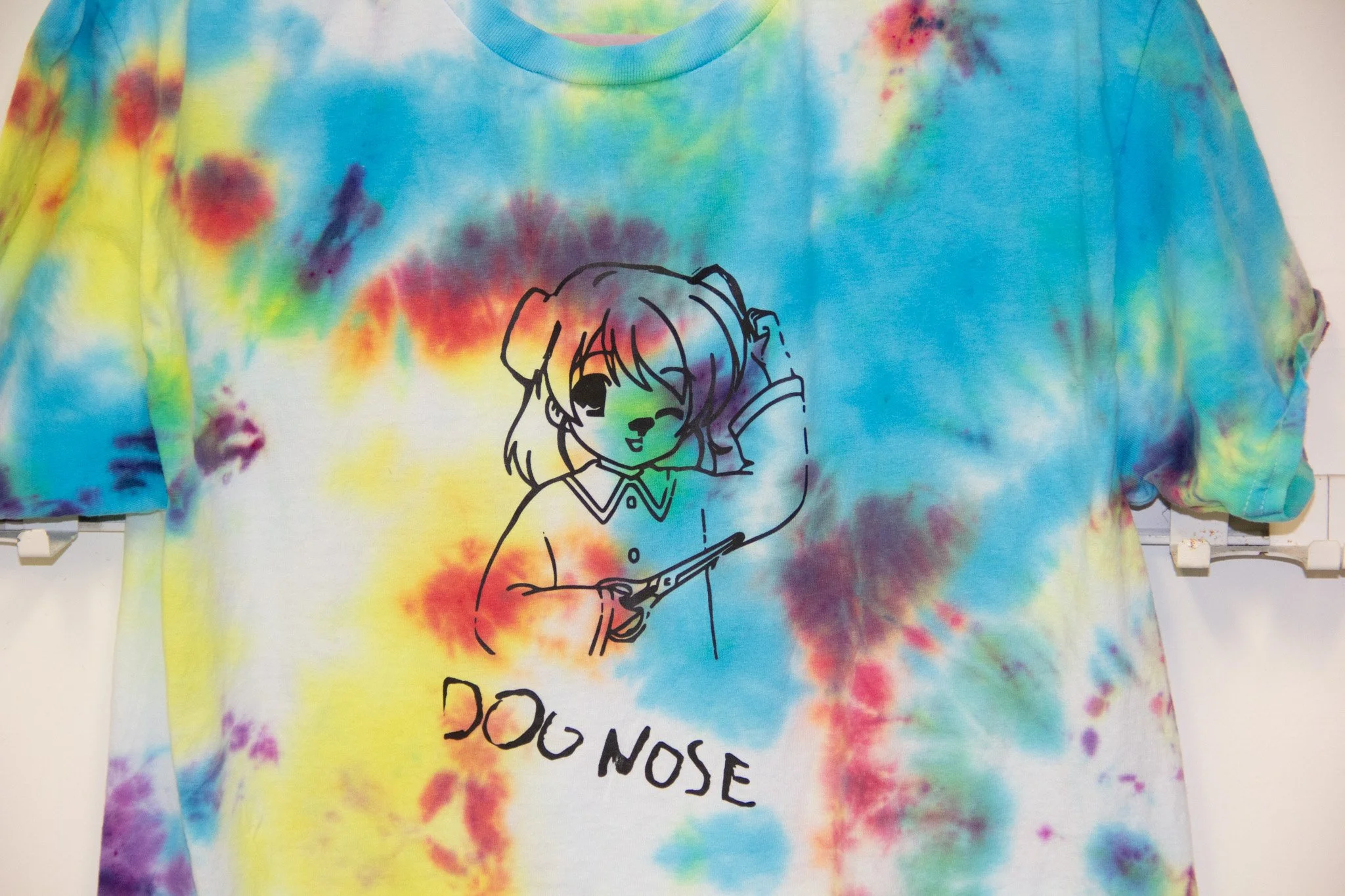 Colorful tie-dye T-shirt featuring a line drawing of a girl with a bow in her hair, playing a guitar, with the words 'DOG NOSE' partially visible underneath.