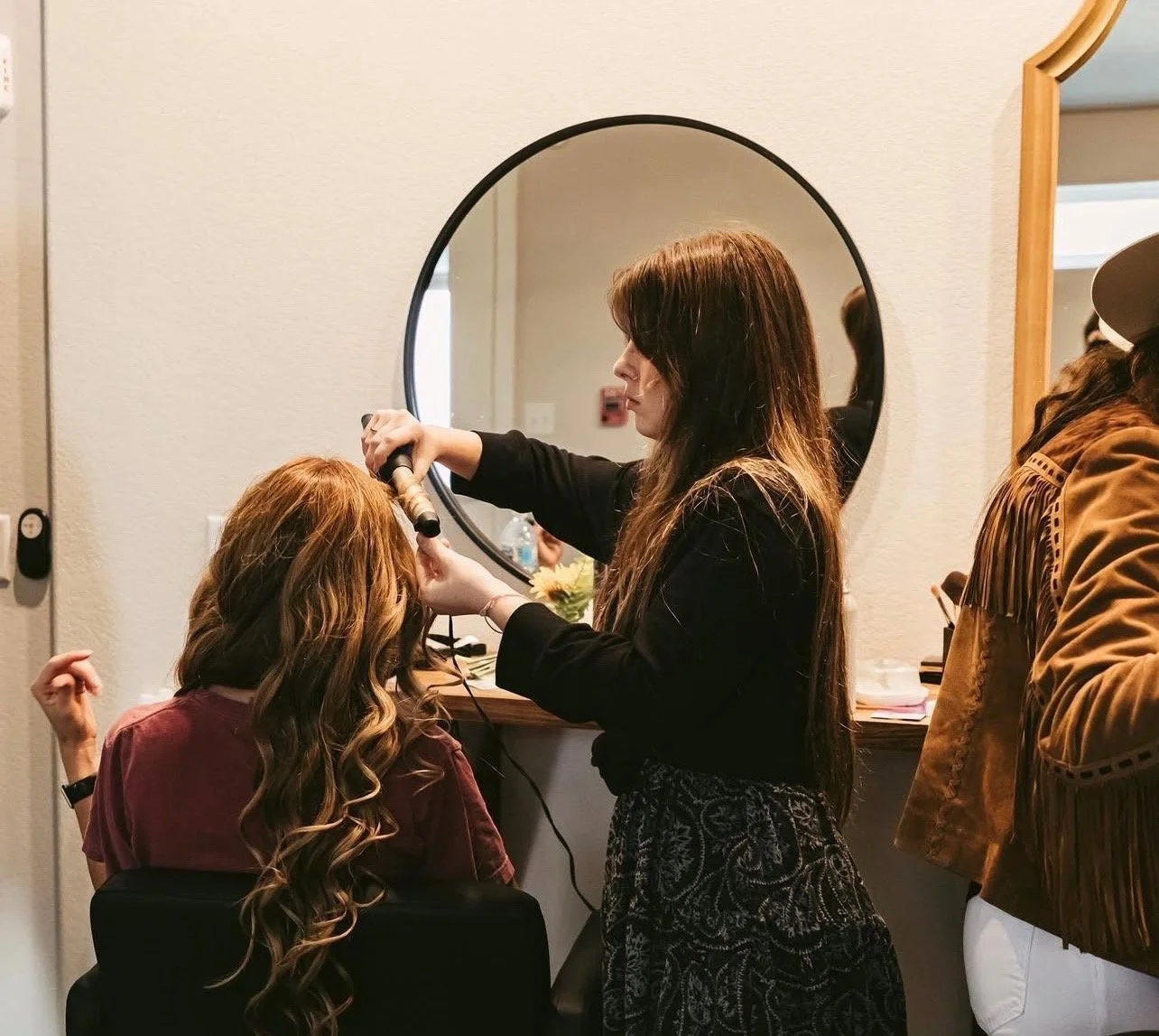 A hairstylist blow-drying a woman's hair while she sits in a chair facing a mirror, with a second woman on the right observing, in a salon setting.