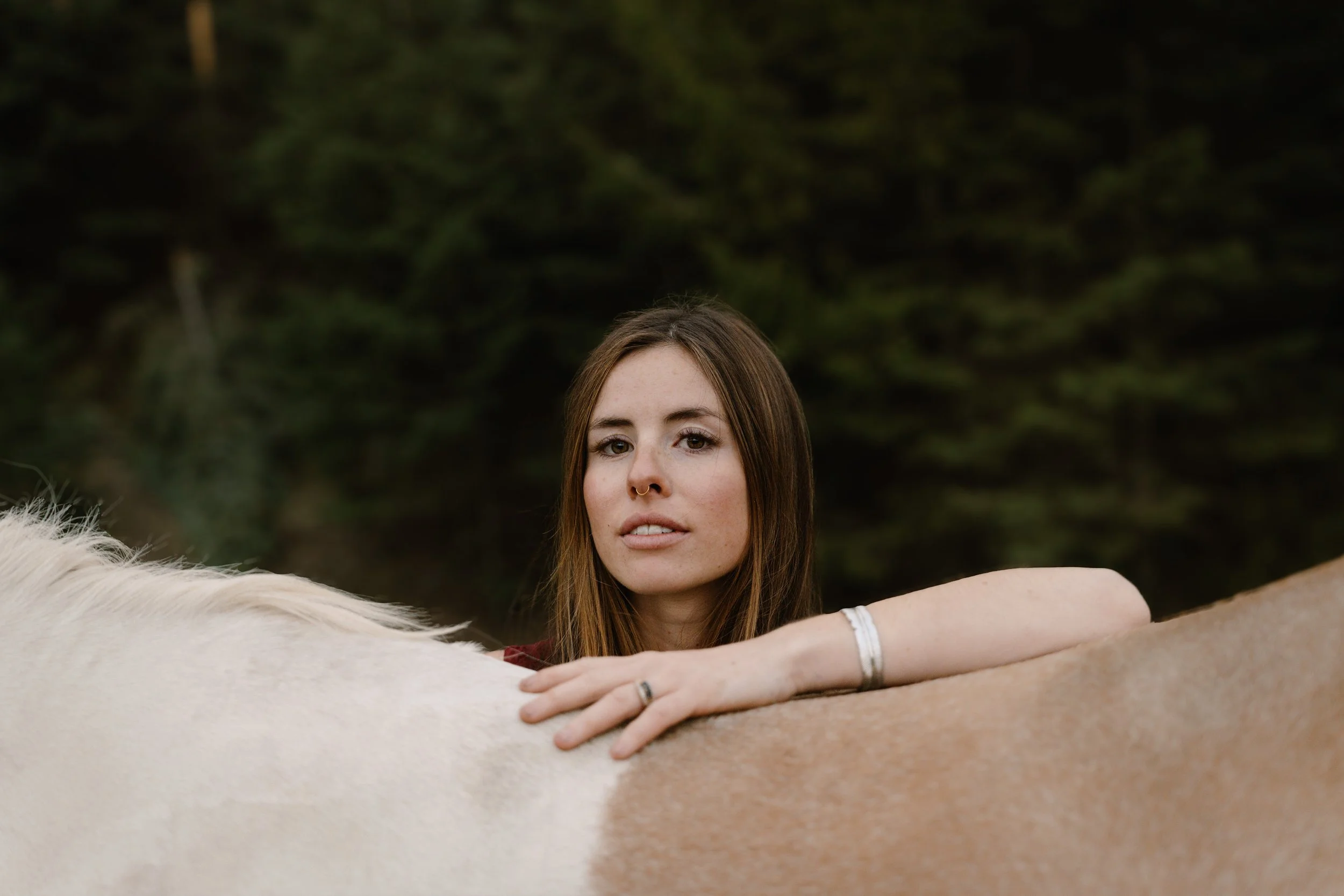 A licensed hair stylist with brown hair and light makeup resting her hand on the back of a white and brown horse outdoors, with a blurred green foliage background.
