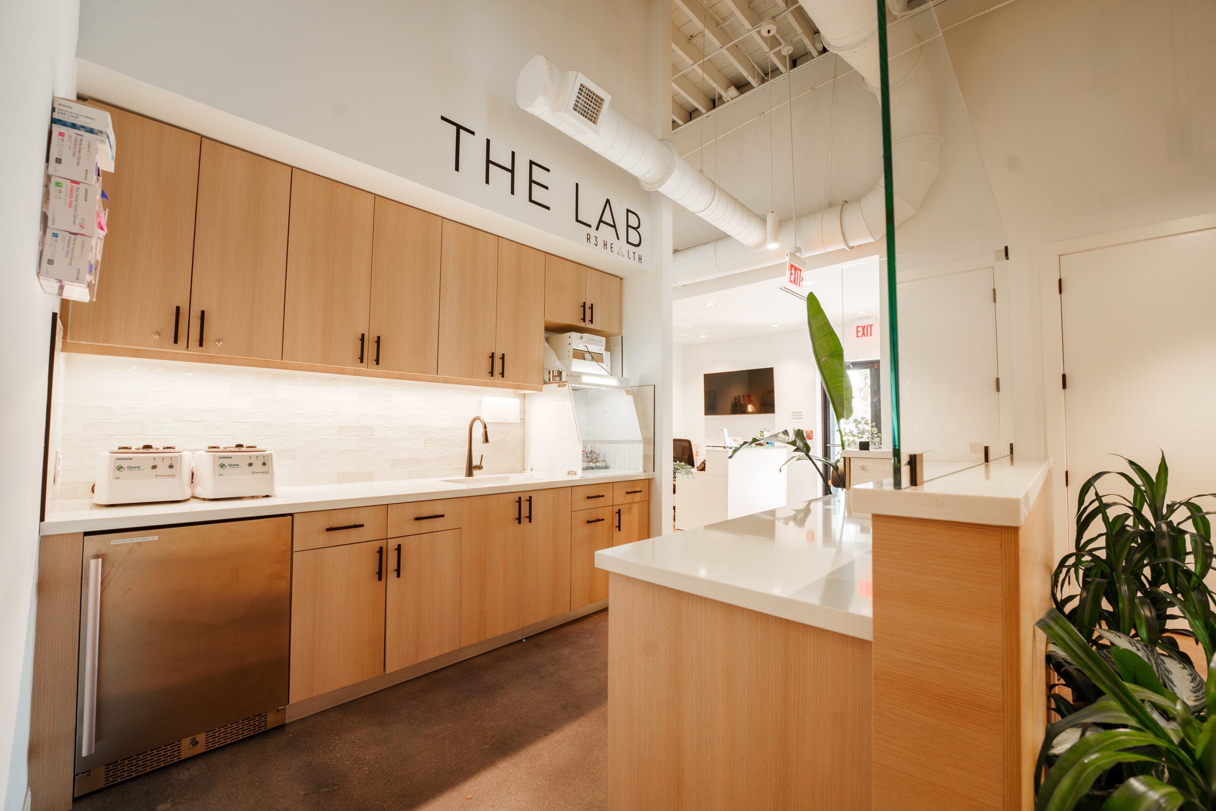 Kitchen area with wooden cabinets, white countertop, sink, two toaster ovens, and a sign that reads 'THE LAB R3 HEALTH' on the wall.