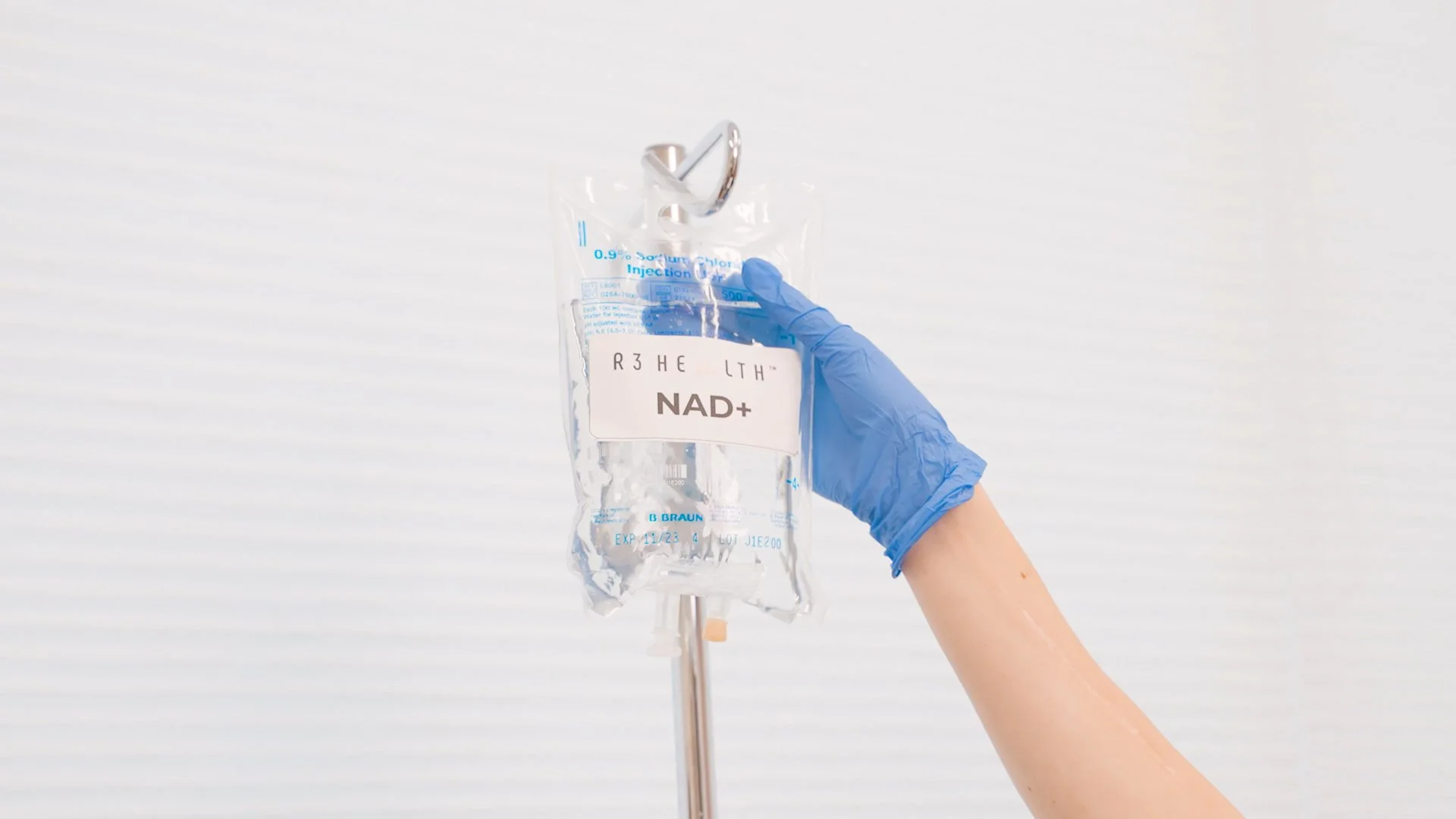 A hand with a blue glove holding an IV bag labeled 'NAD+' against a plain, light background.