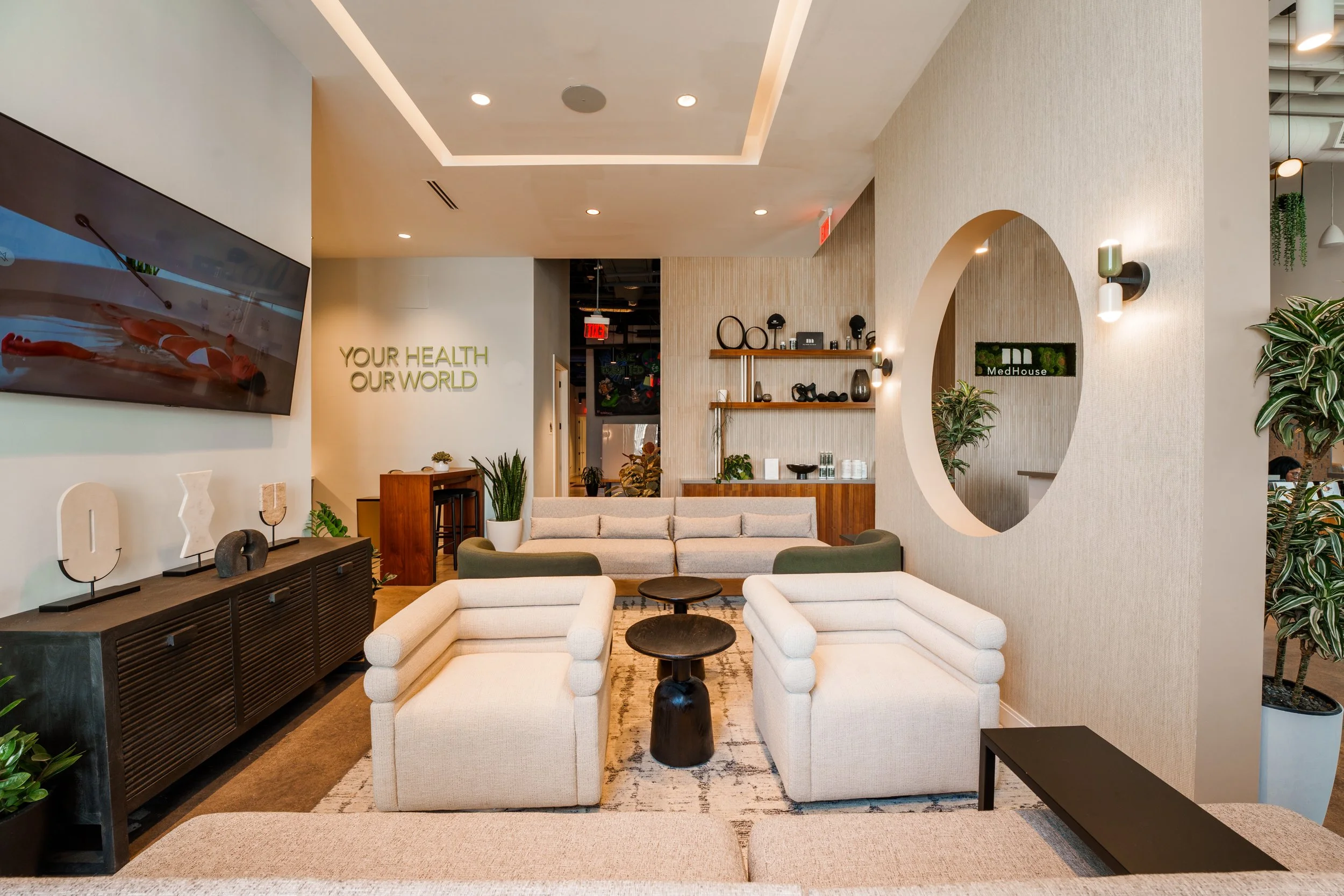 Modern wellness center lobby with beige and green seating, plants, wall art, and a large TV, emphasizing health and relaxation.