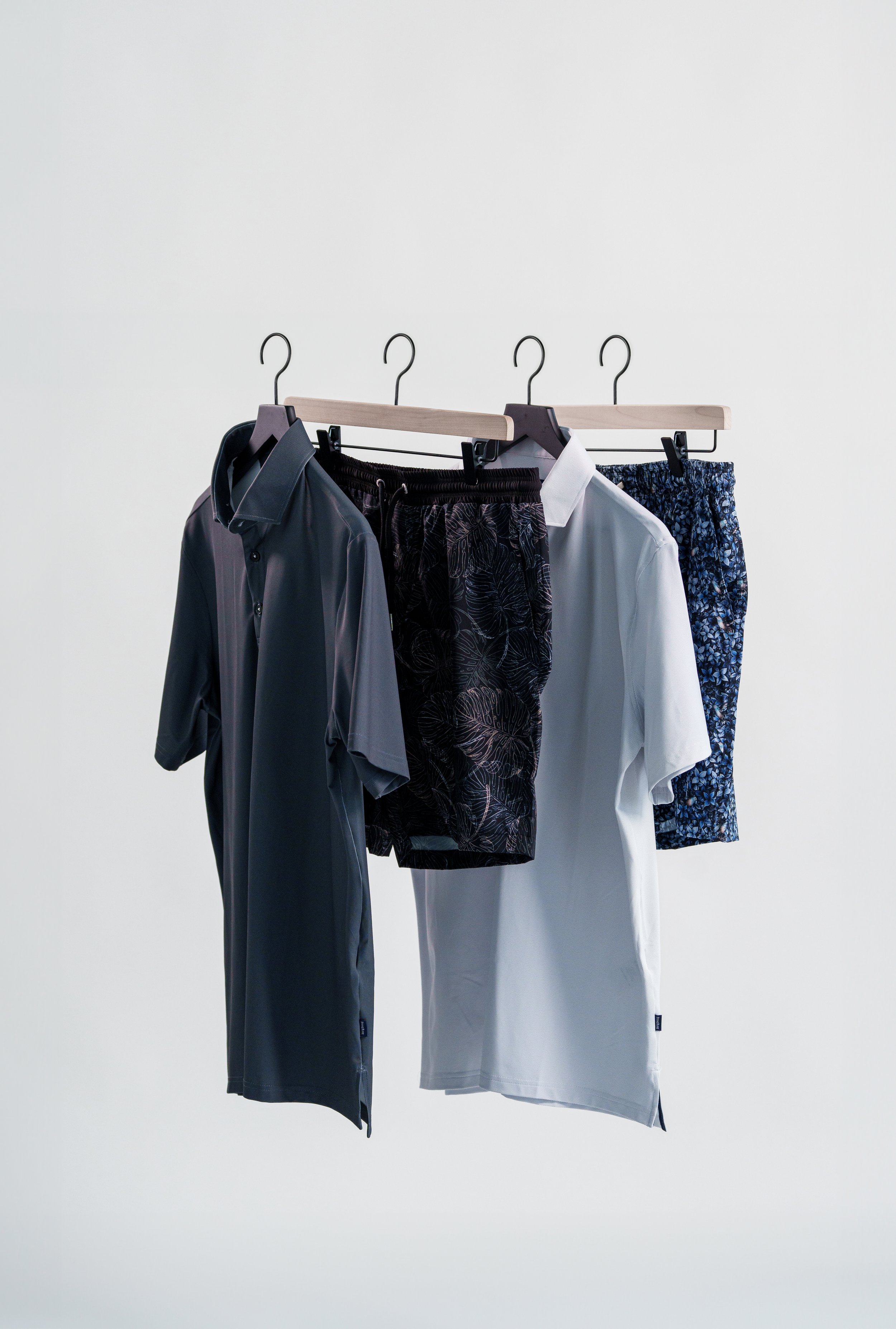Clothing hanging on a rack, including a dark gray short-sleeve shirt, black patterned shorts, white shirt, and blue patterned shorts, against a plain white background.