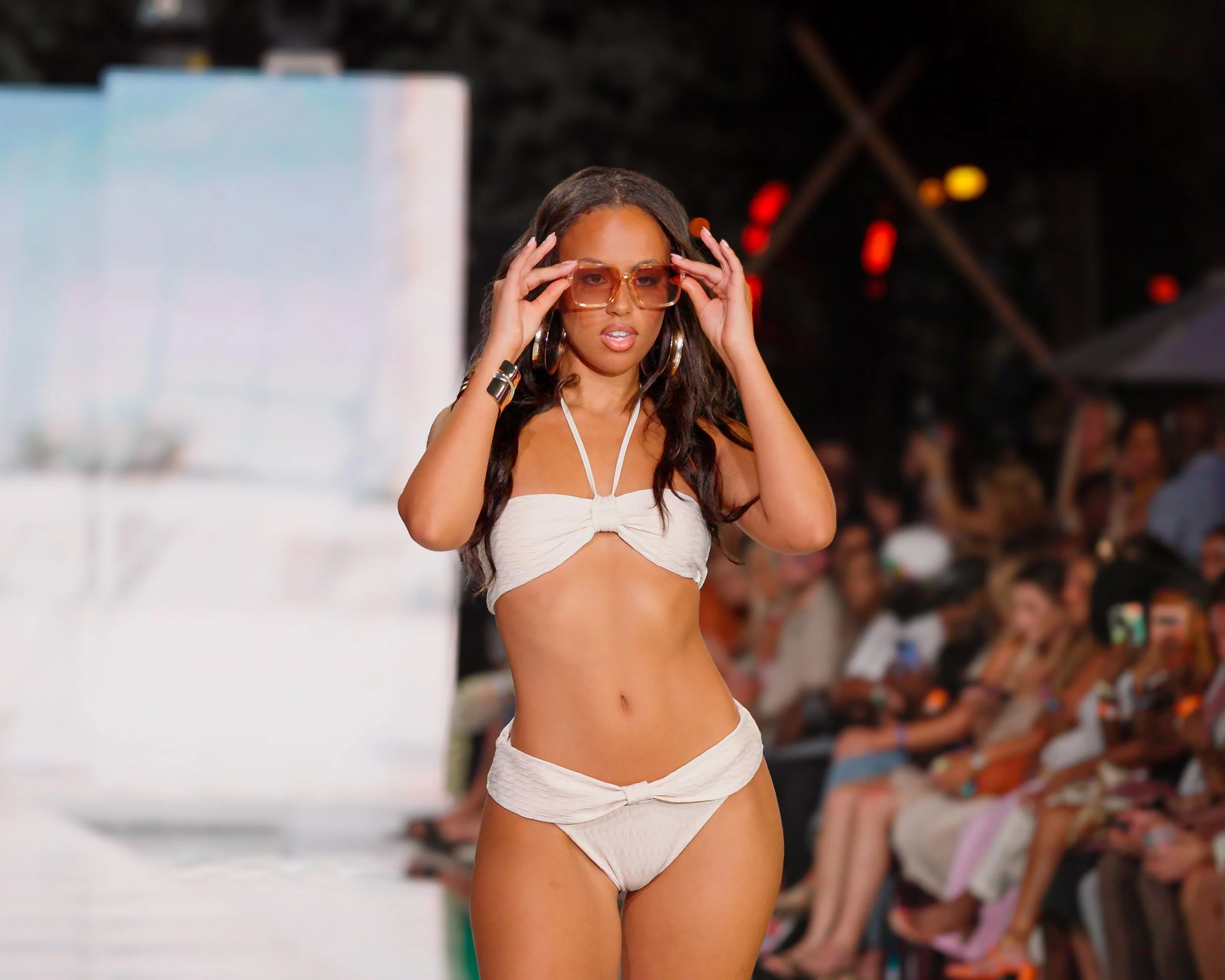 A woman on a runway wearing a white bikini with large sunglasses, surrounded by an audience watching the fashion show at night.