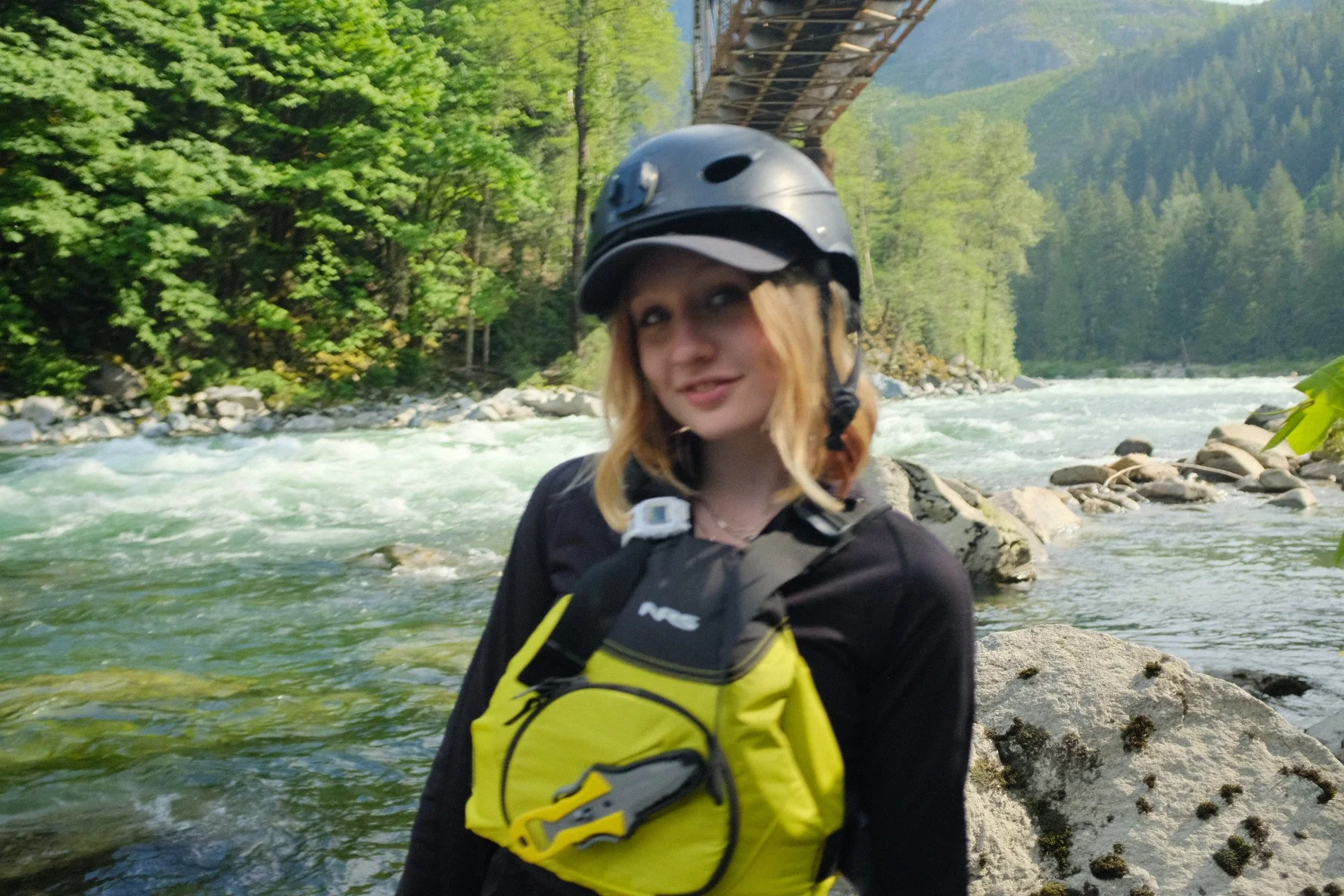 Woman wearing a black helmet and yellow life jacket beside a rushing mountain river beneath a bridge