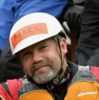 Bearded man wearing a white helmet labeled “MARK” and an orange-and-black motorcycle jacket