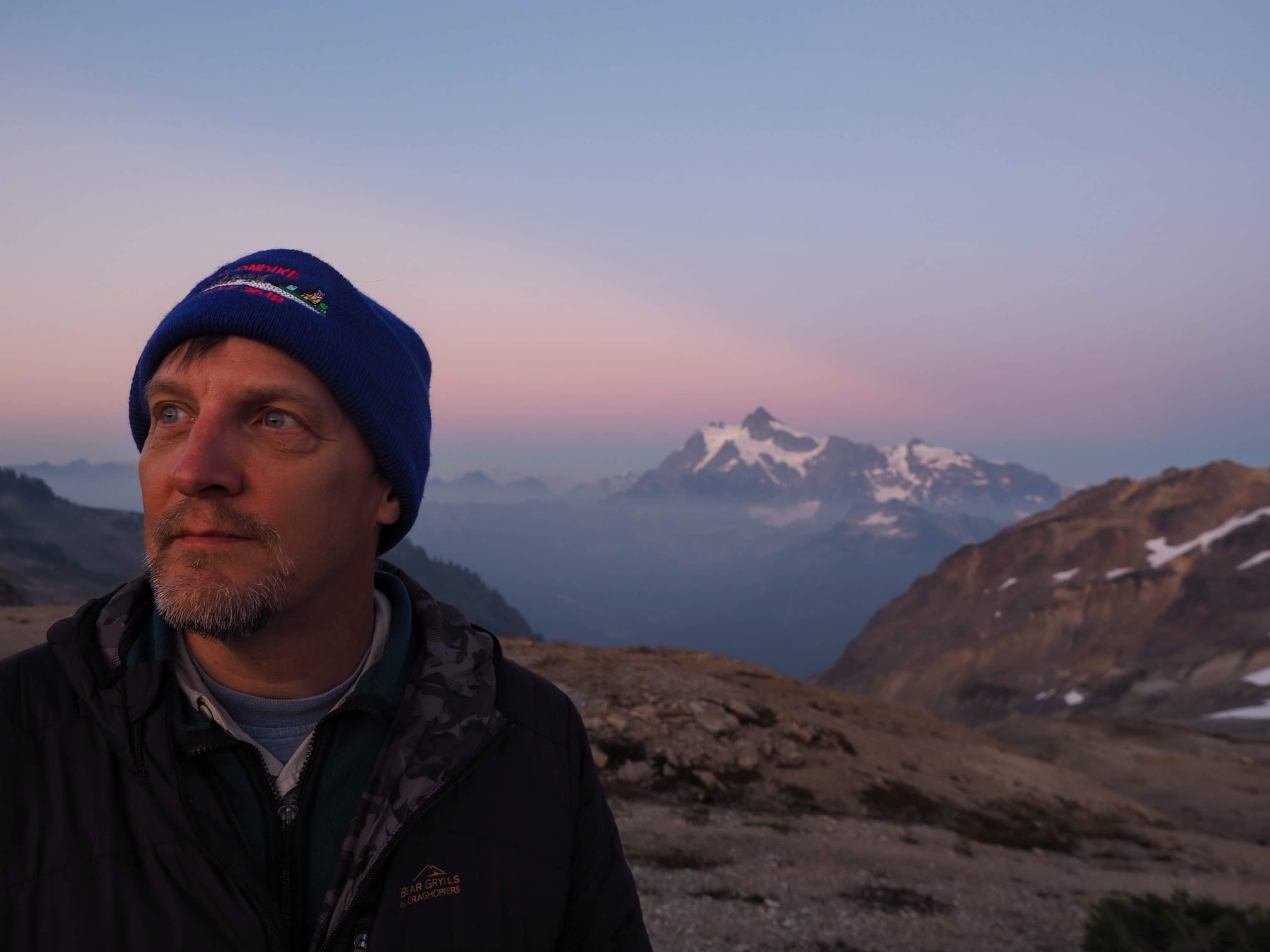 Hiker wearing a blue beanie and outdoor jacket with a snow-capped mountain range at sunset