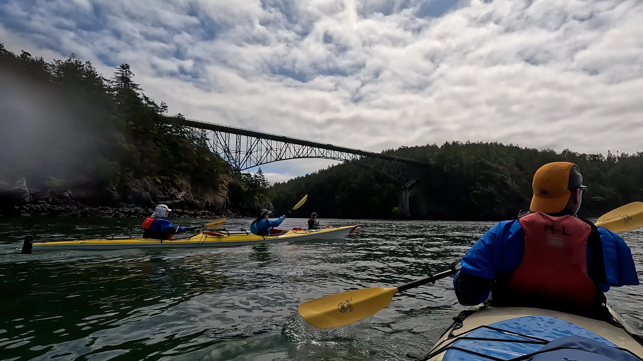 Two people kayaking in a red kayak on a body of water, wearing life jackets and hats, with rocky cliffs and mountains in the background.