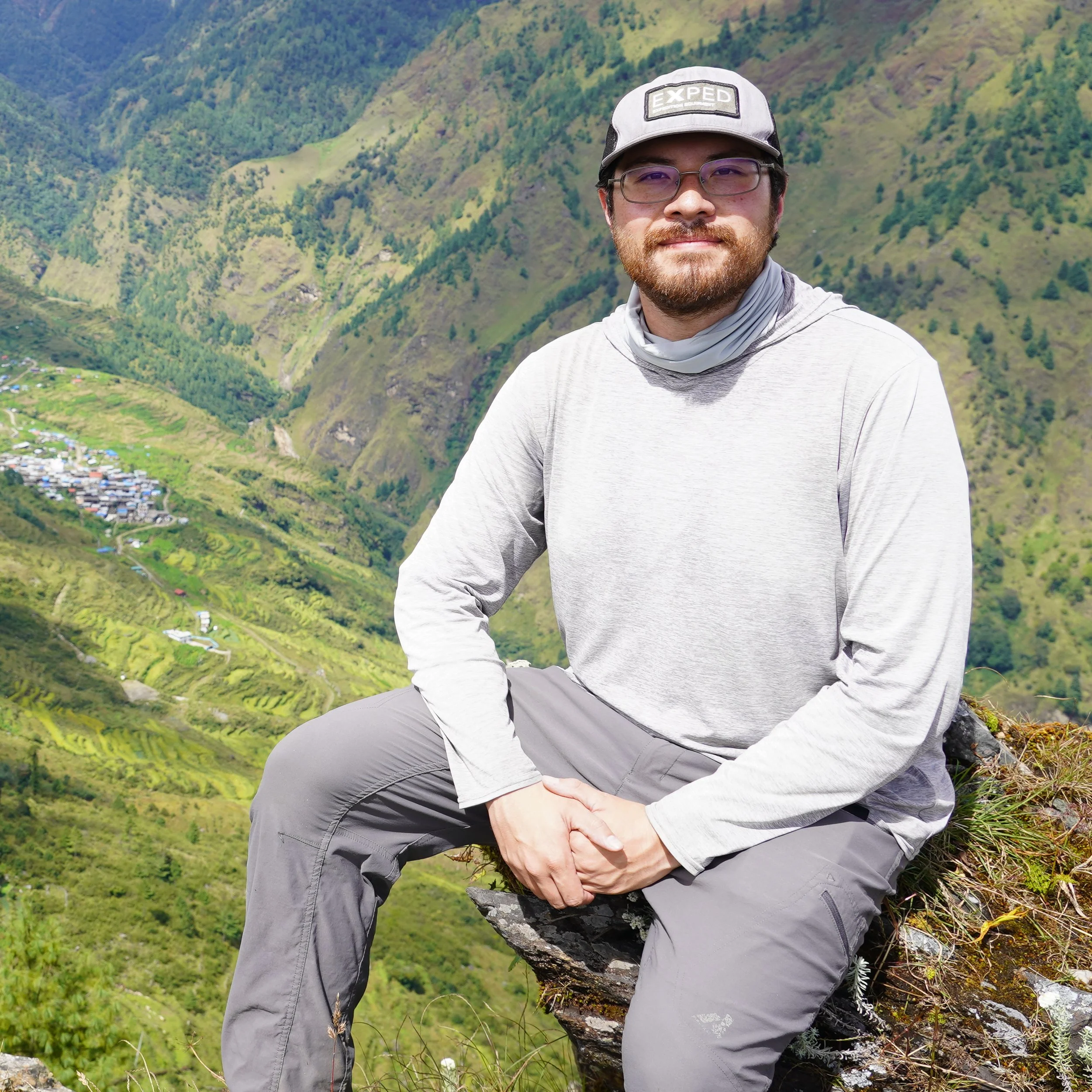 Traveler wearing glasses and a cap sits on a rocky ledge overlooking a lush mountain valley and village.