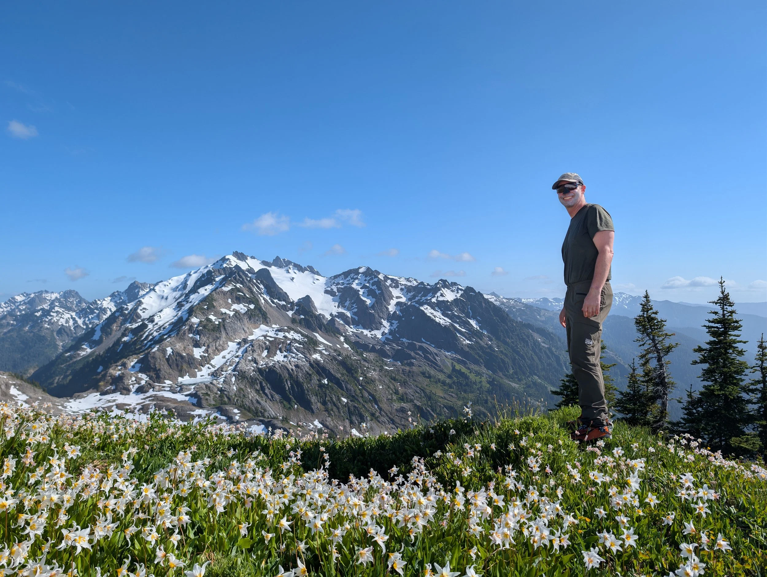 Hiker standing in a wildflower meadow overlooking snow-capped mountain peaks under a clear blue sky