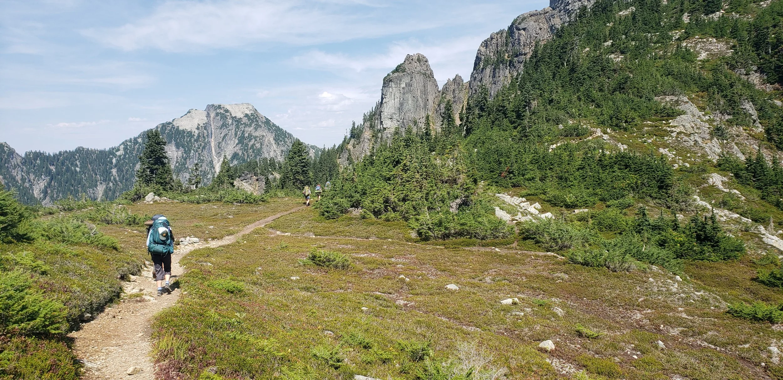 Two hikers overlooking a scenic mountain valley with snow-capped peaks and a winding river on a cloudy day.