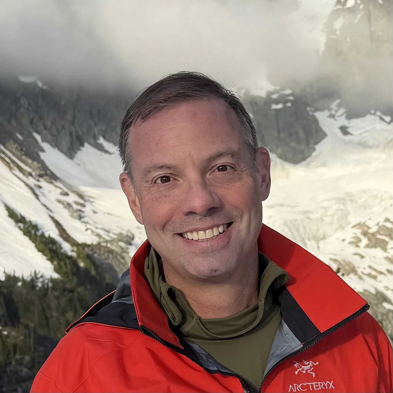 Smiling man in a red Arc'teryx jacket taking a selfie in front of snowy mountain slopes.