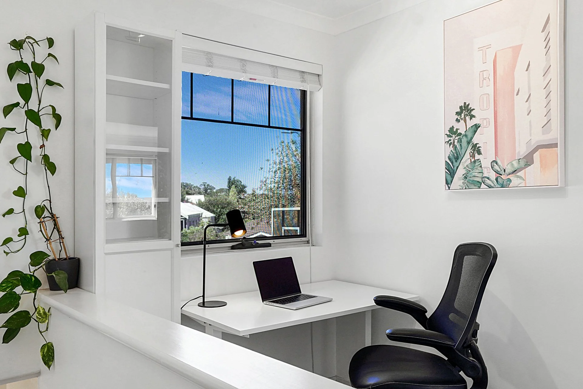 Modern white office with a window view of blue sky and trees, a laptop on a white desk, black office chair, desk lamp, and a framed tropical plant art on the wall.