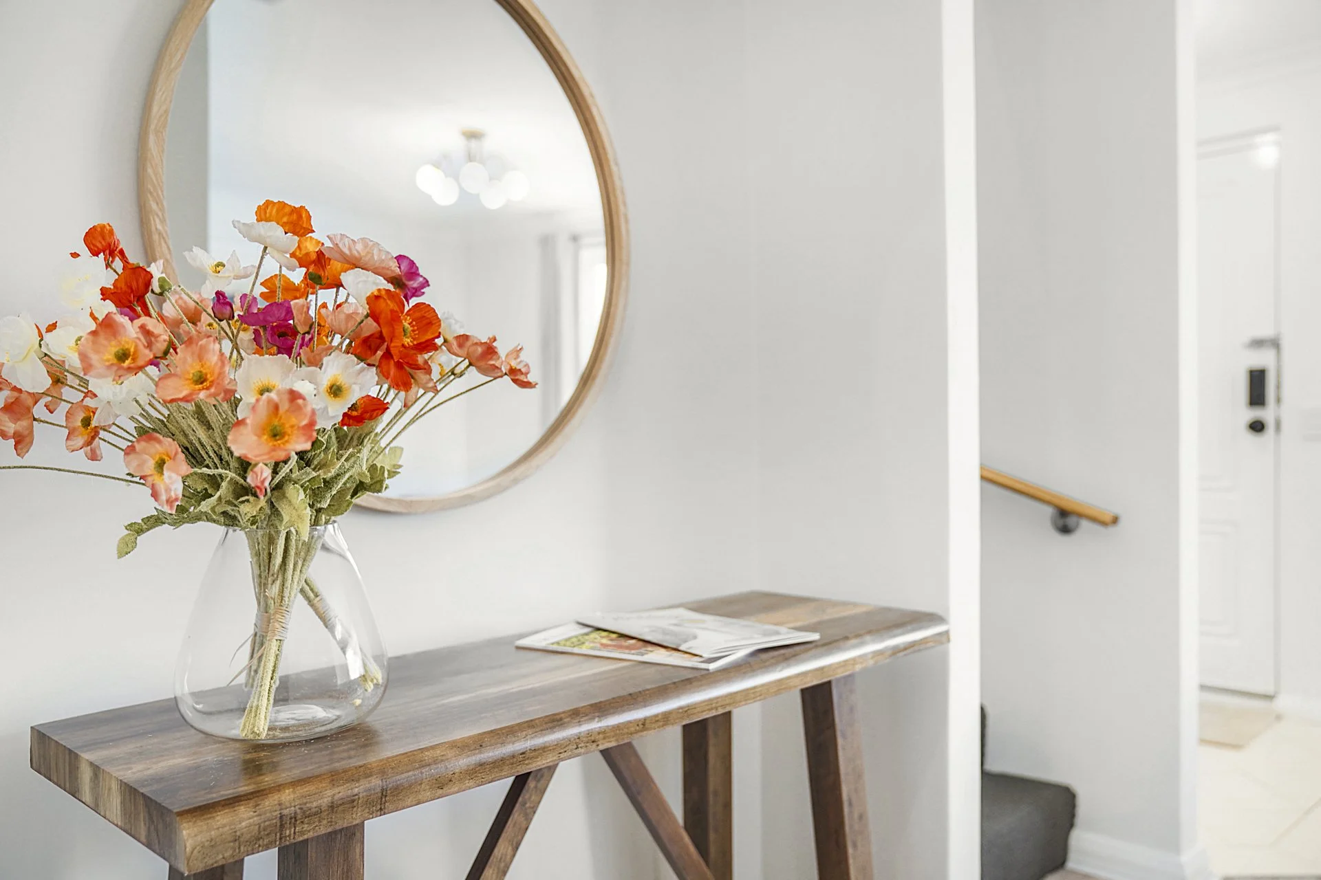 A glass vase with a bouquet of colorful flowers, including orange, pink, and white blossoms, sits on a wooden table in front of a round mirror on a white wall. Sunlight and ceiling lights are reflected in the mirror. HostYou