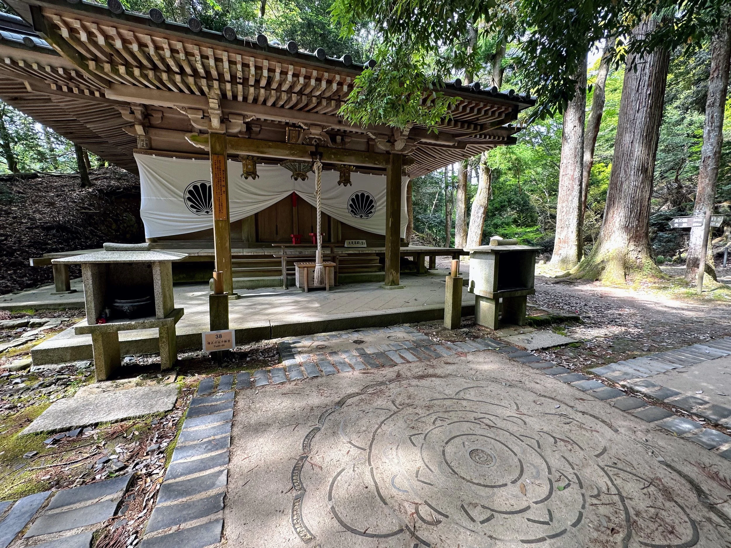 A small traditional Japanese shrine in a forested area, with wooden structure and white curtains decorated with black symbols, surrounded by trees and stone pathways.