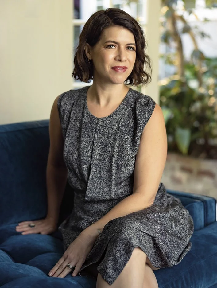 A woman with short brown hair sitting on a blue couch, wearing a sleeveless gray dress, in a bright room with plants in the background.