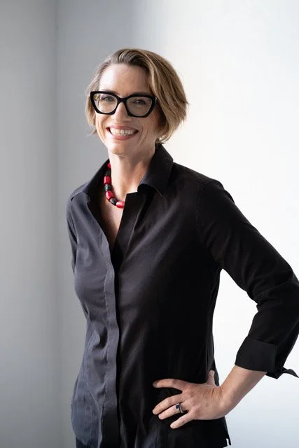 Woman with short hair, wearing glasses and a black shirt with a colorful necklace, standing indoors against a white wall