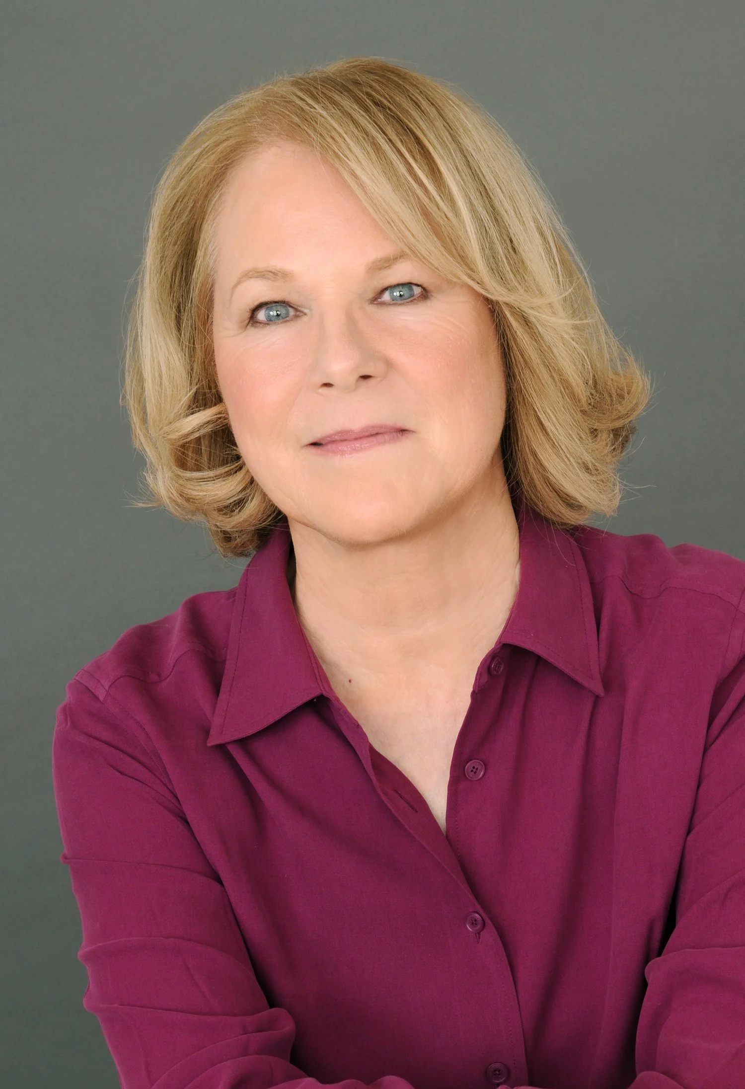 A woman with blonde hair and blue eyes wearing a maroon shirt posing against a gray background.
