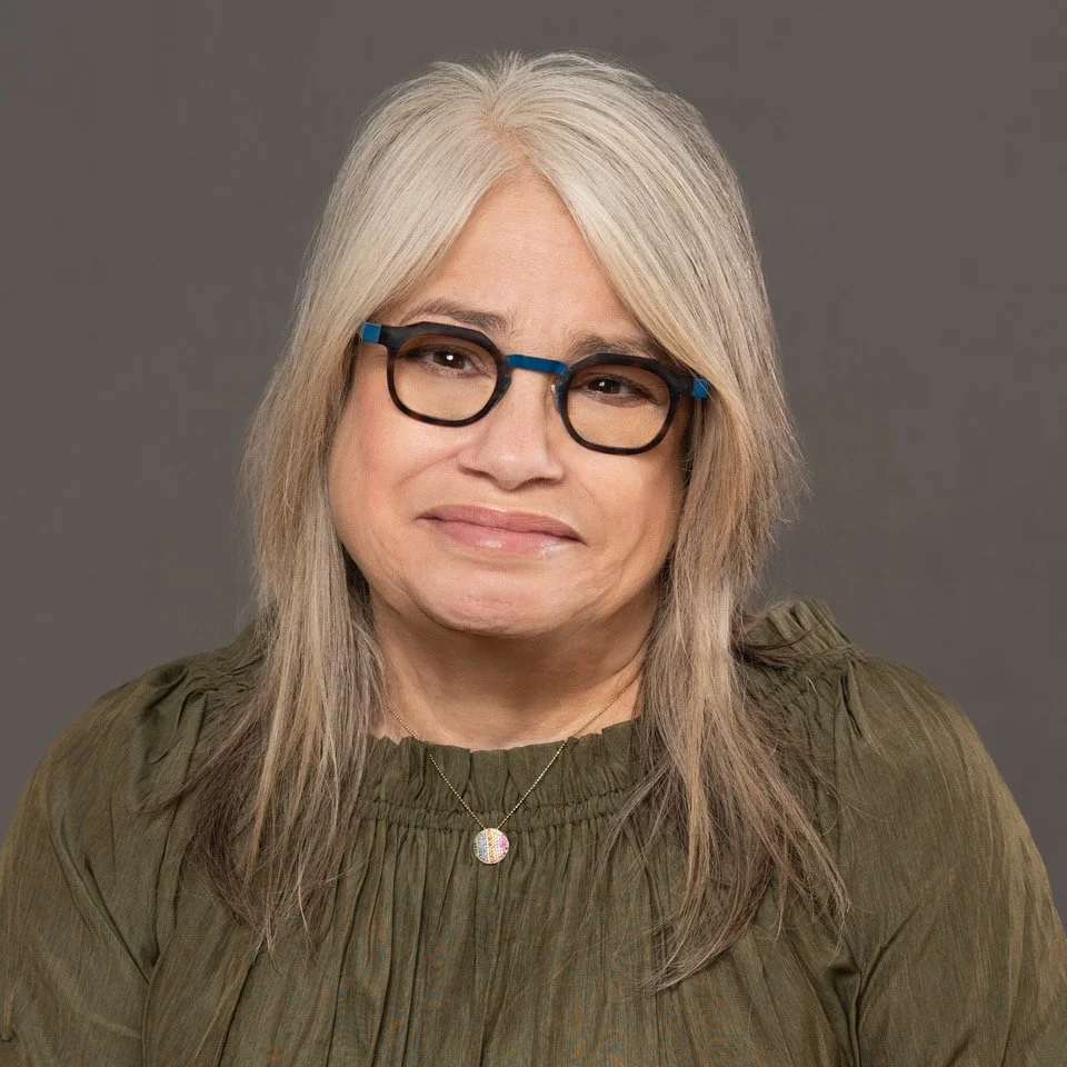 A woman with shoulder-length gray hair, glasses with blue accents, wearing a green top, and a gold necklace with a colorful pendant, standing against a gray background.