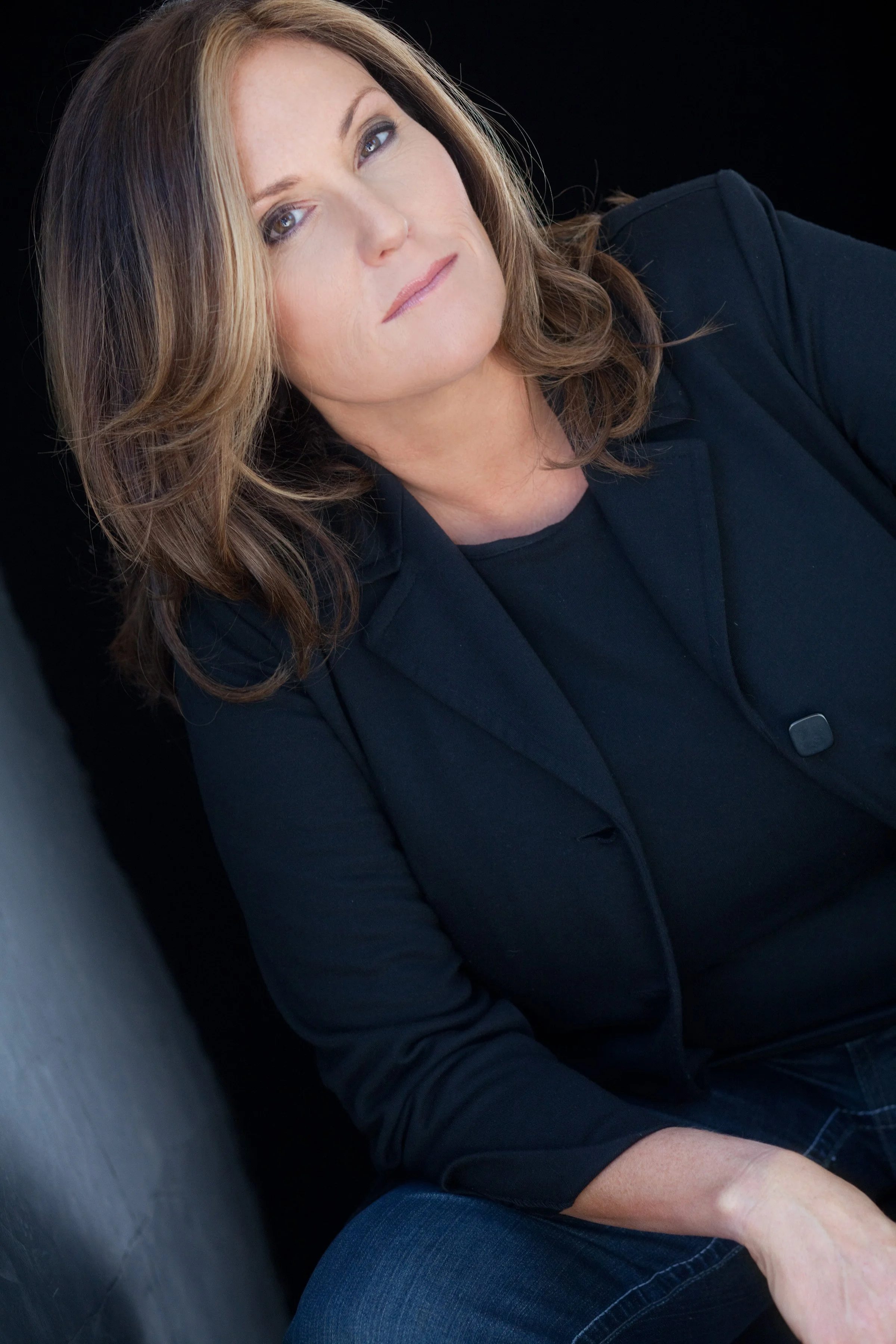A woman with shoulder-length brown hair and blue eyes, dressed in a black blazer and black shirt, sitting against a dark background.