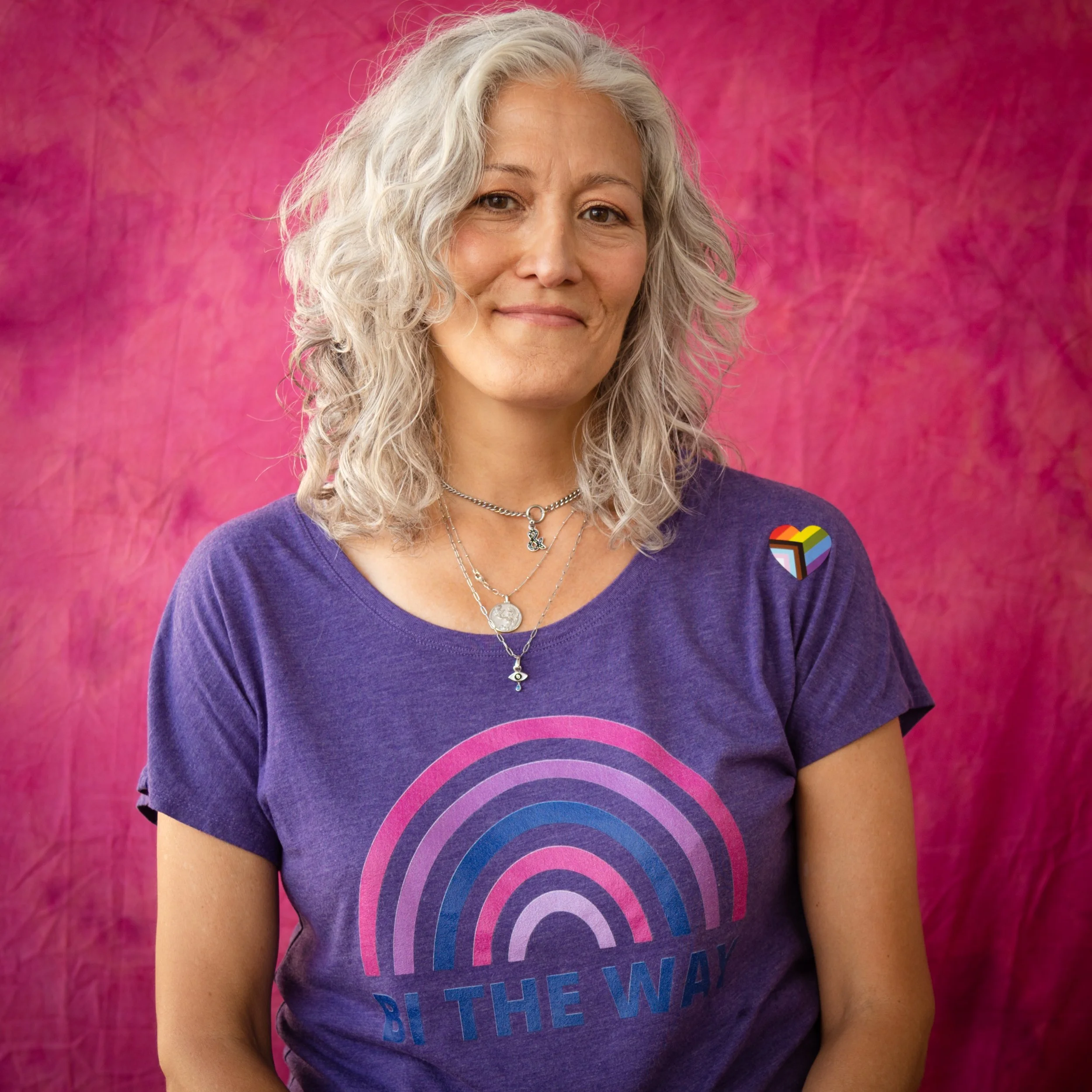 A woman with curly gray hair, wearing a purple t-shirt with a rainbow design and the words 'BI THE WAY', standing against a pink background with a rainbow pride pin on her shoulder.