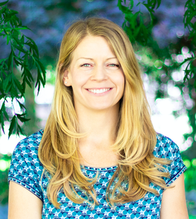A smiling woman with long blonde hair standing outdoors with green trees in the background.
