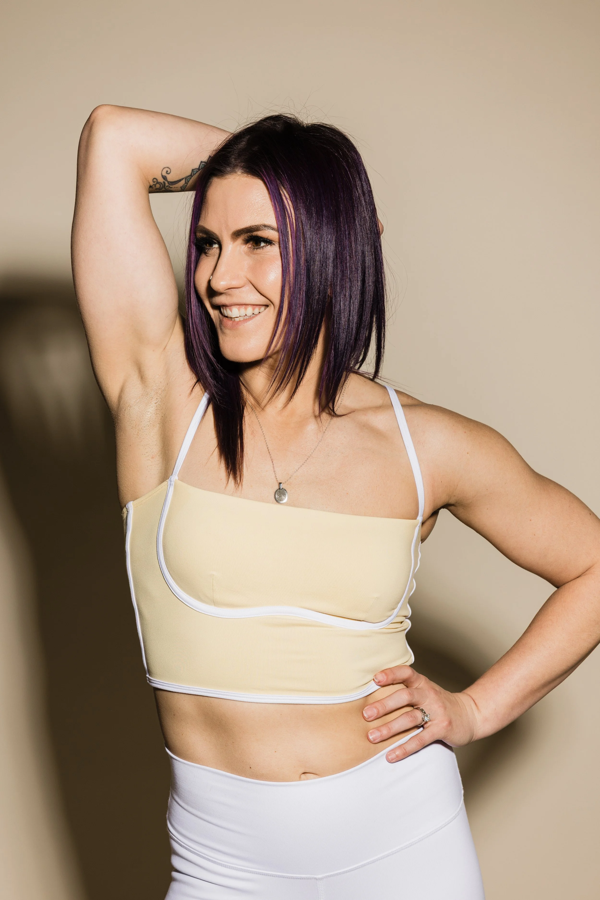A woman with purple hair smiling, wearing a yellow crop top and white pants, standing with one hand on her hip and the other behind her head.