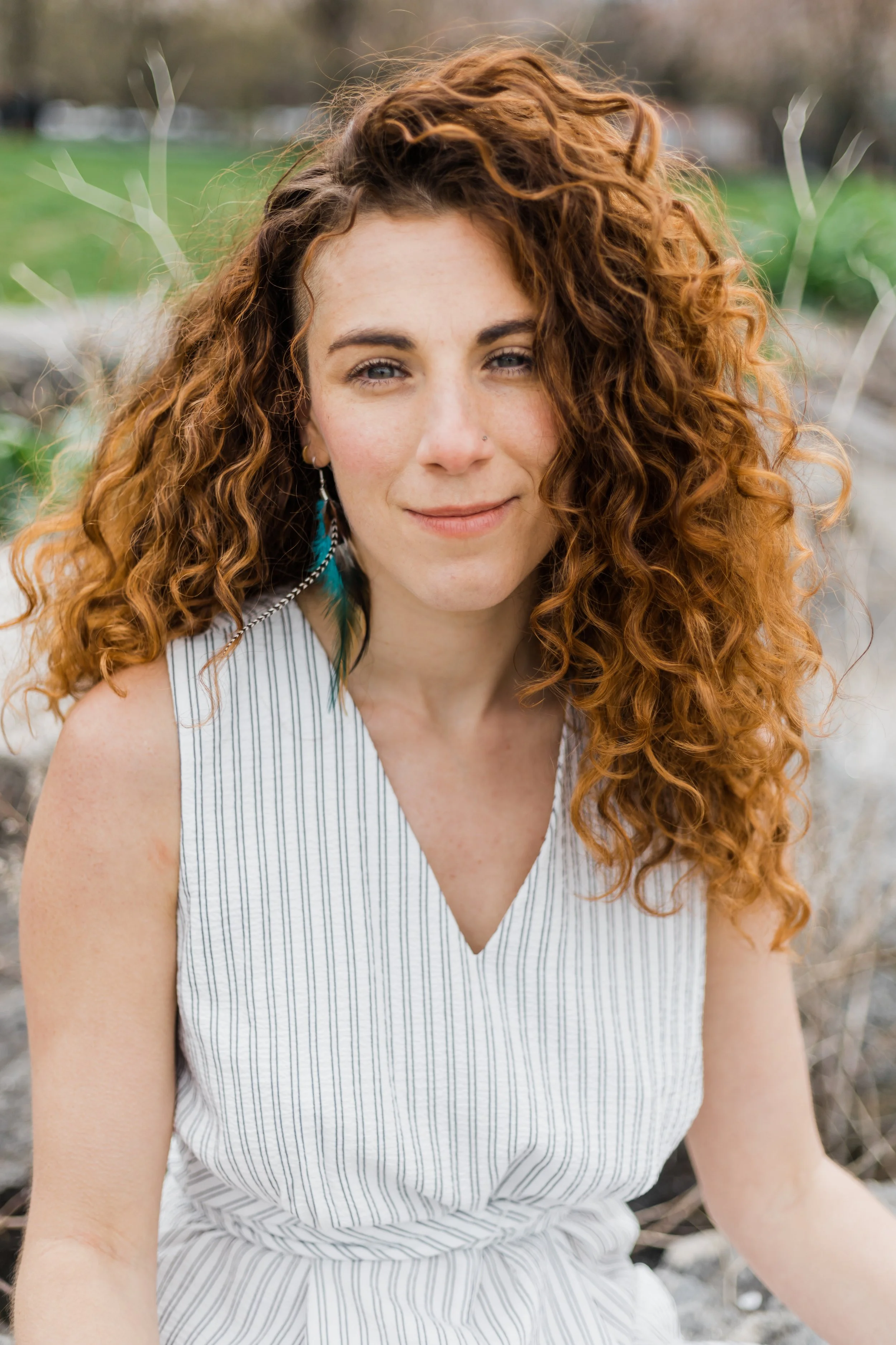 A woman with curly red hair wearing a sleeveless, striped top, standing outdoors with a blurred natural background.