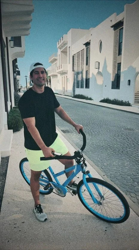 A young man wearing a black T-shirt, neon yellow shorts, and a white cap standing on a sidewalk with his blue bicycle, in a sunny urban area with white buildings.
