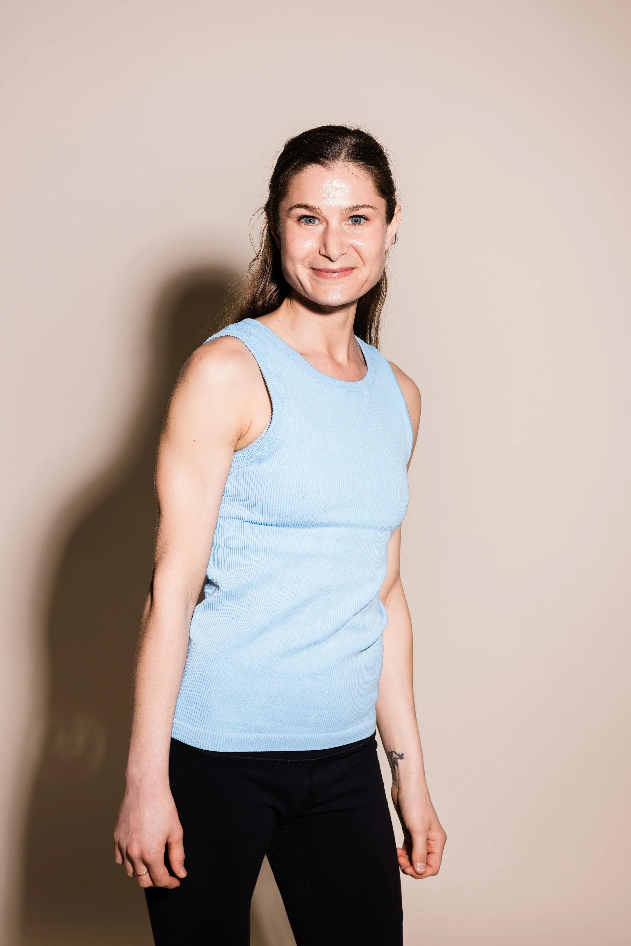 A woman with long brown hair, wearing a light blue sleeveless top and black pants, standing against a beige background with a slight shadow.