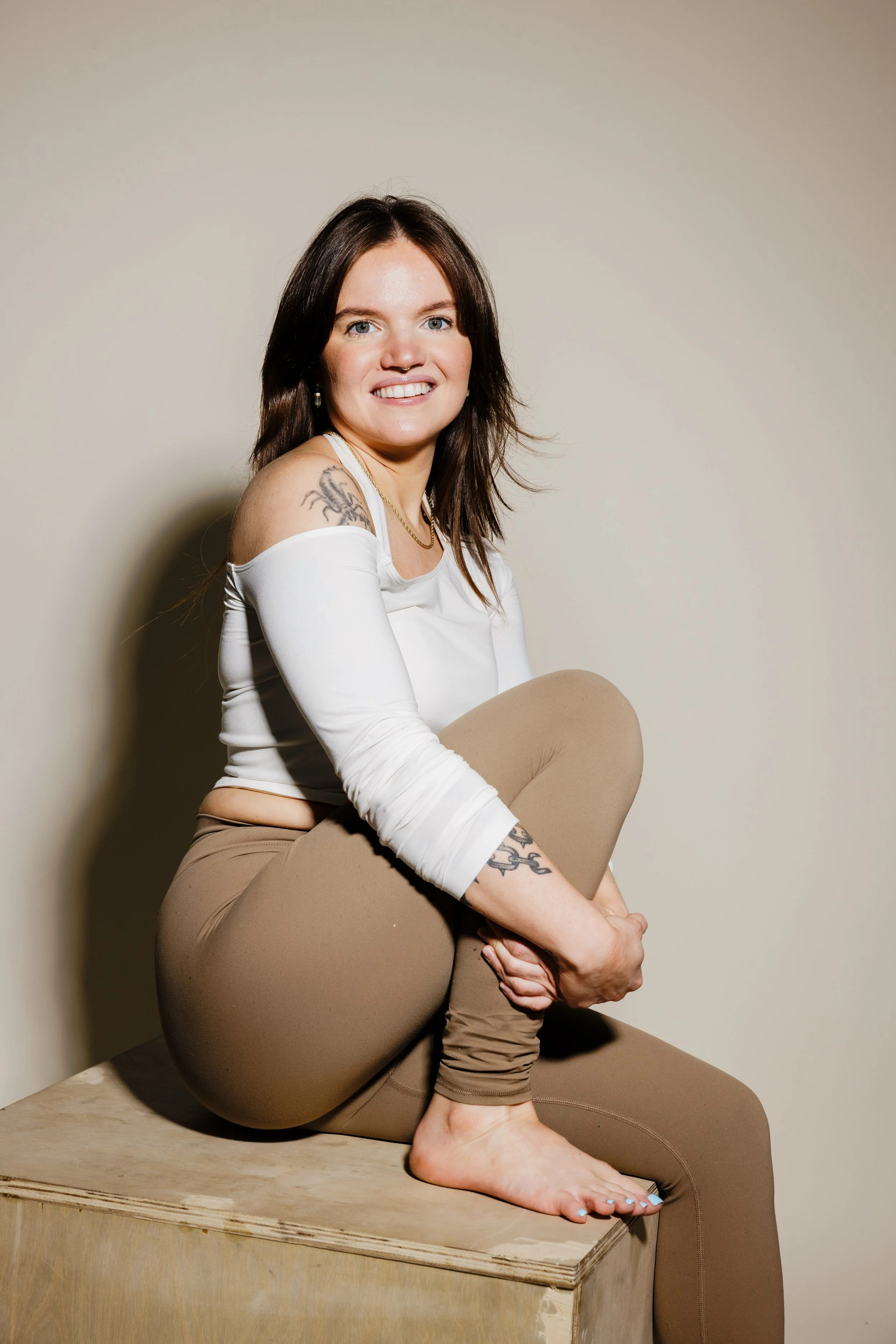 A woman with brown hair, tattoos, and light skin, sitting on a wooden surface against a plain background, smiling at the camera, in casual clothing.