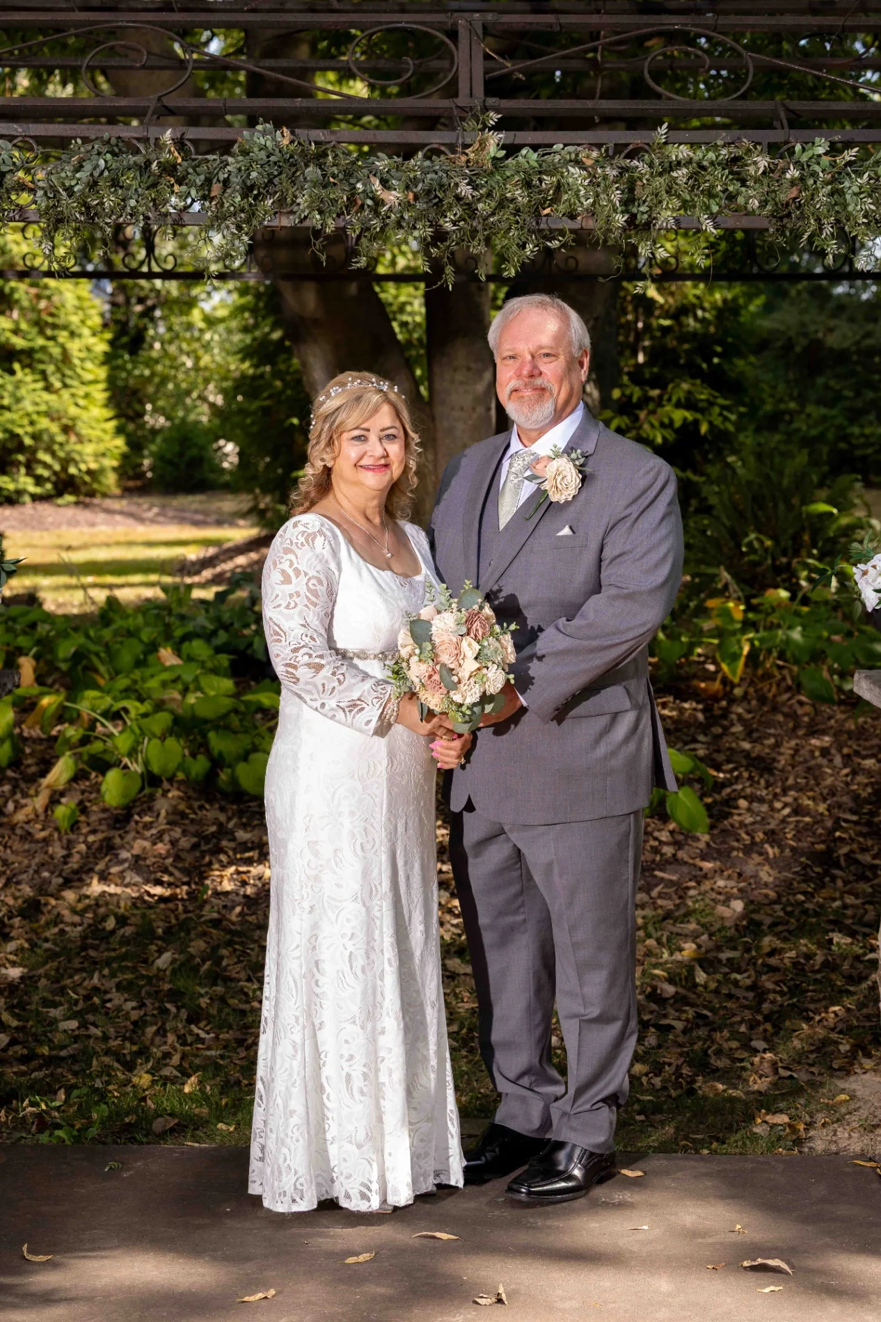 A couple in formal attire stands outdoors; the woman wears a white lace dress and holds a bouquet, while the man wears a gray suit with a boutonniere. Greenery surrounds them.