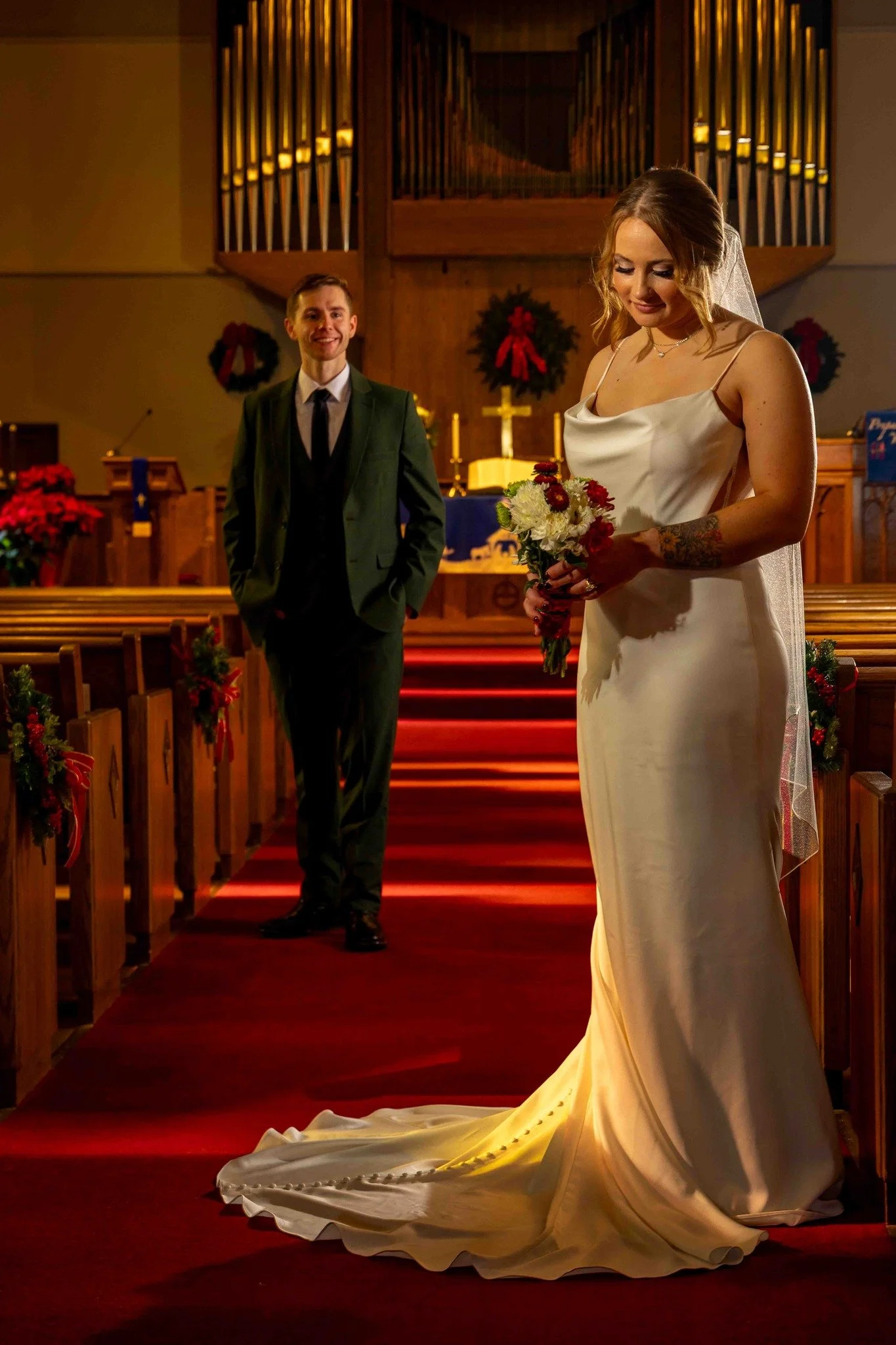 A bride in a white gown holds a bouquet and stands at the front of a church while a groom in a dark suit stands smiling in the background.