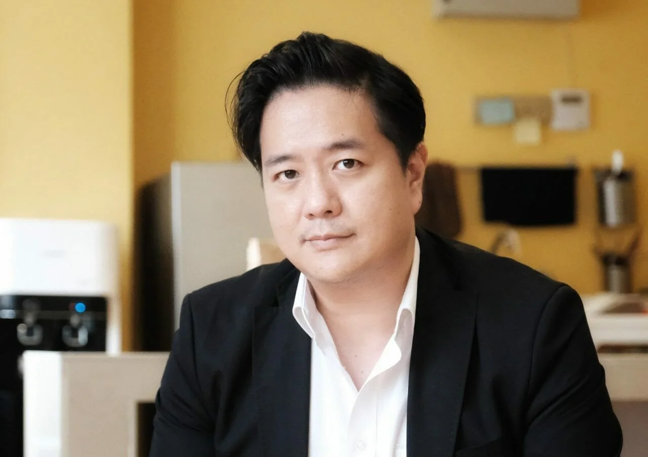 A man with dark hair is wearing a black suit jacket over a white shirt, seated in an office setting with a yellow wall and various office equipment in the background.
